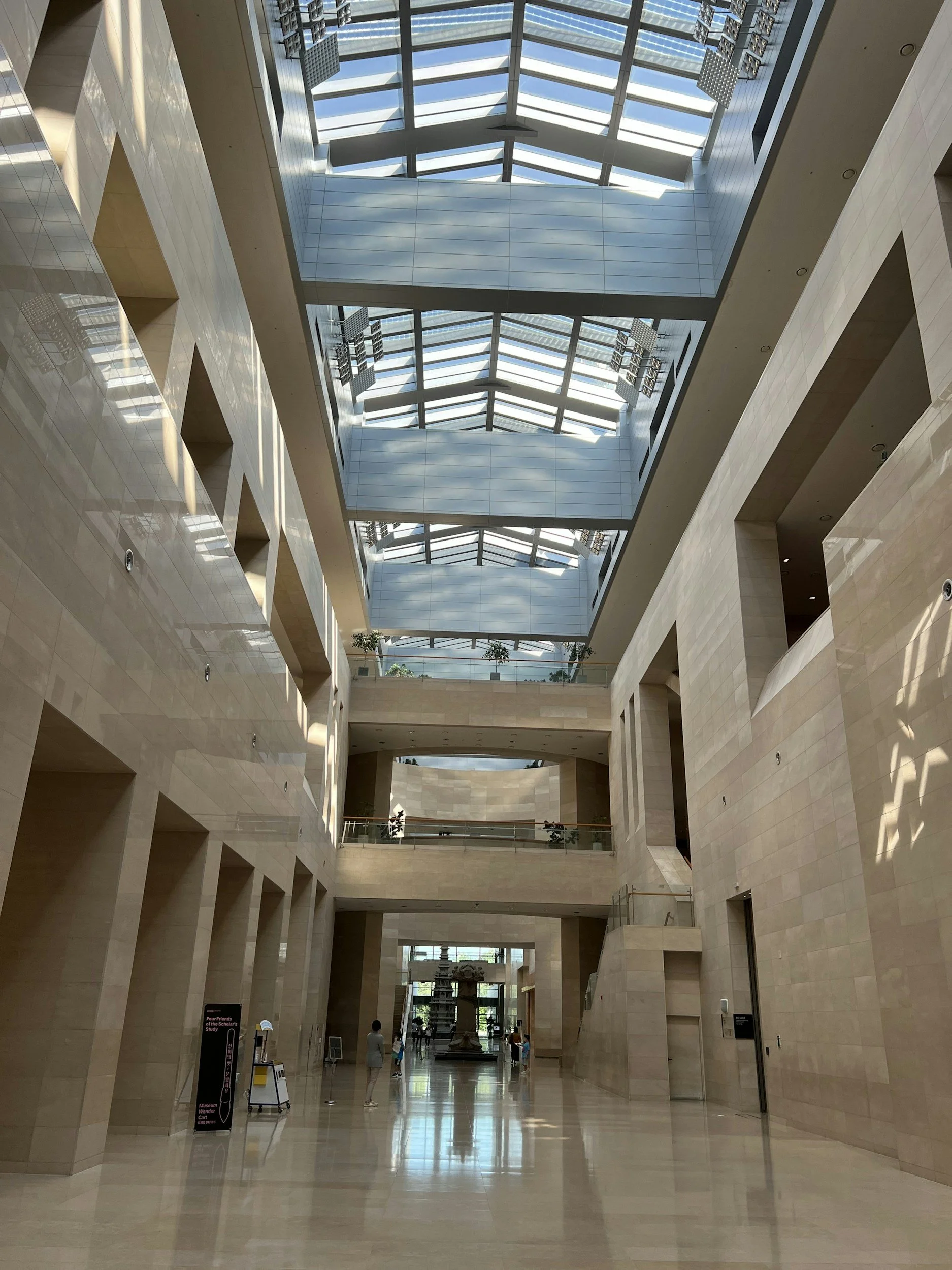 Interior view of a spacious modern building with high ceilings and large glass skylights, beige tiled walls, and several people walking in the distance.