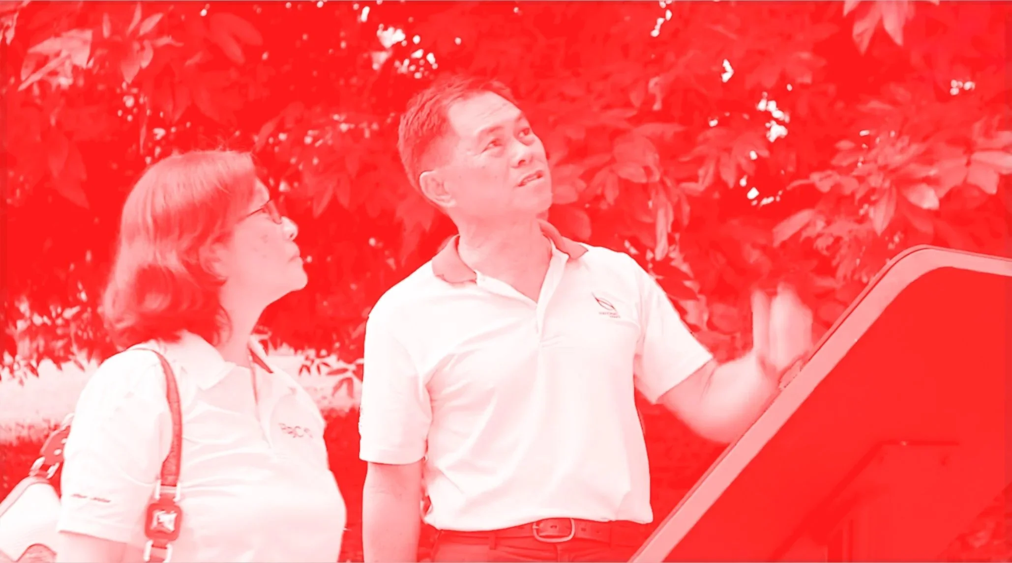A man and a woman standing outdoors with trees behind them. The man is pointing at something on a red kiosk or display, and the woman is looking at the same direction.
