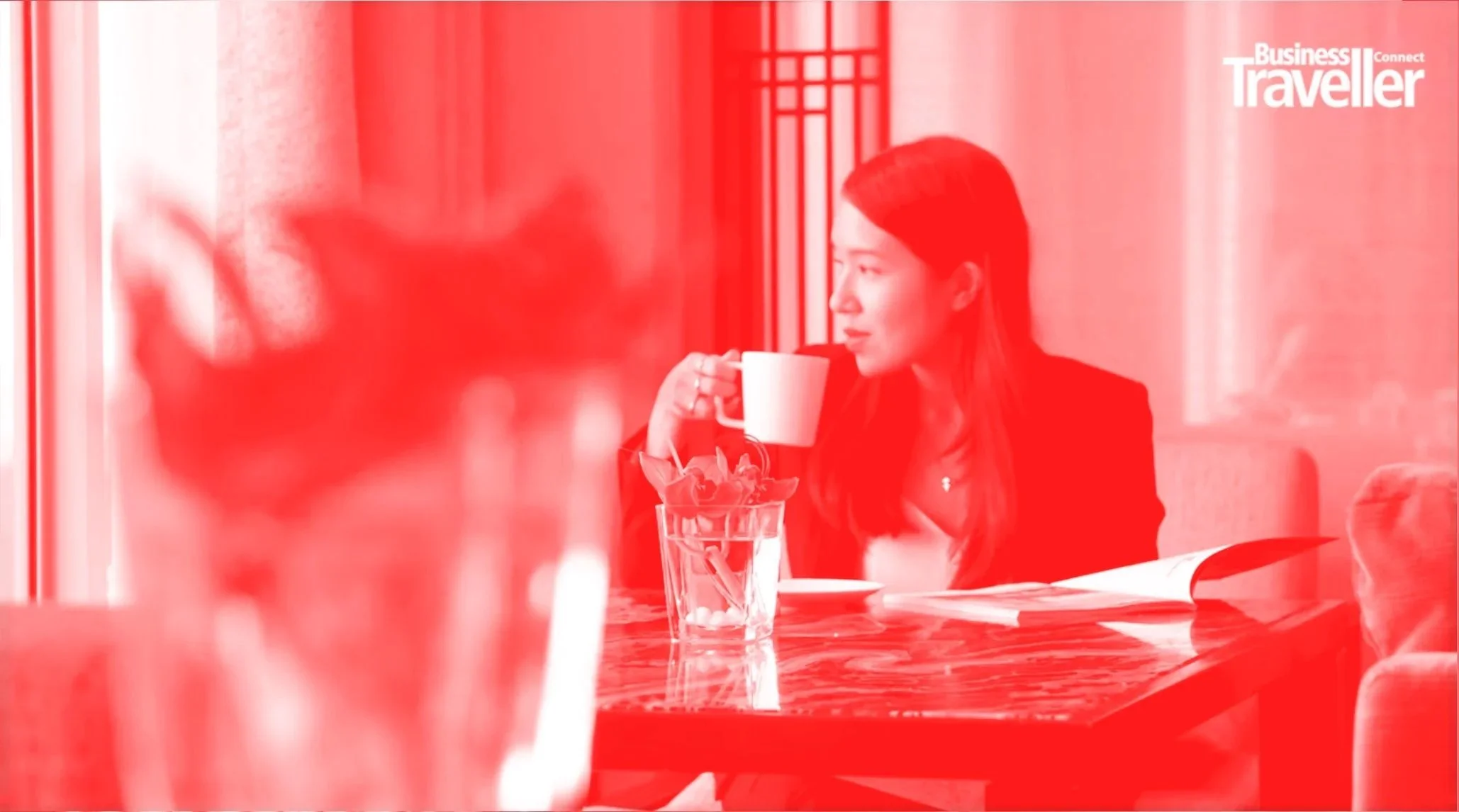 A woman sitting at a table with a glass of water, a notepad, and a magazine, holding a mug and looking out the window in a cafe.