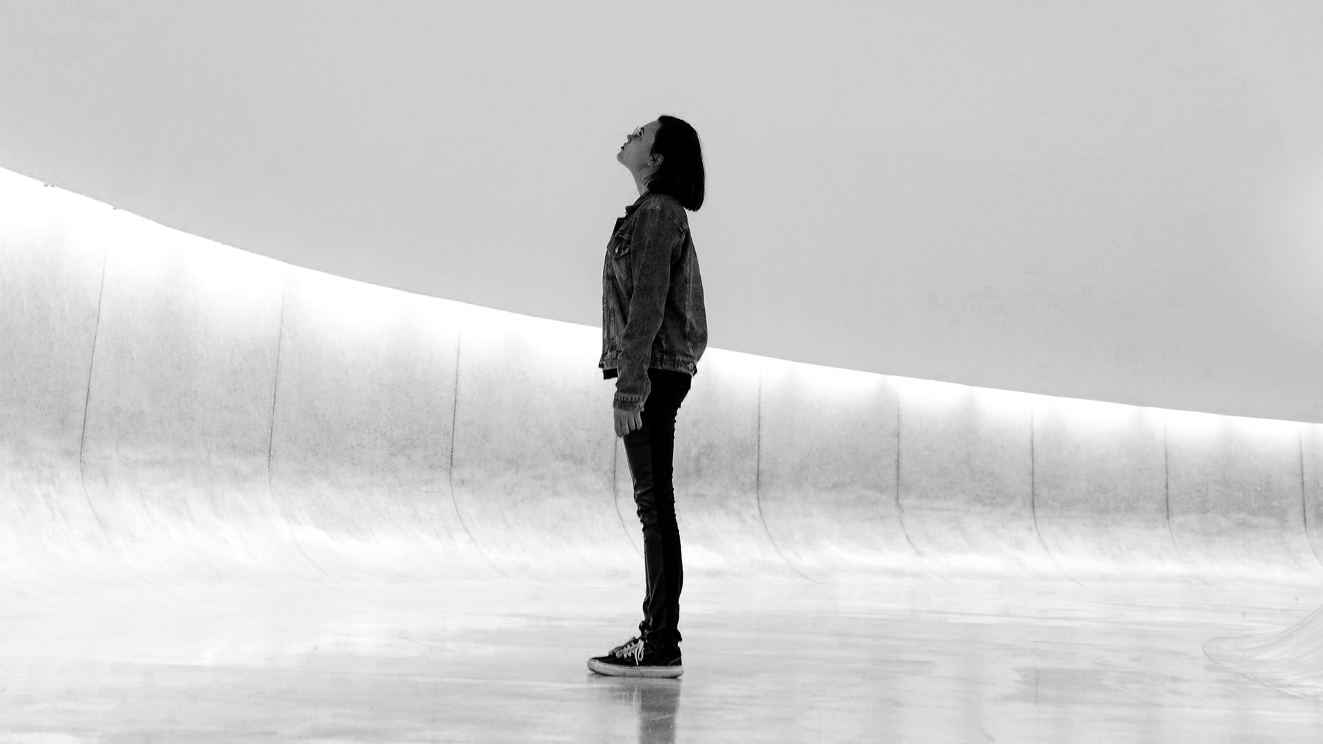 A woman standing indoors, facing upward, wearing a denim jacket, pants, and sneakers, in a spacious, modern, minimalist space with curved architectural features.