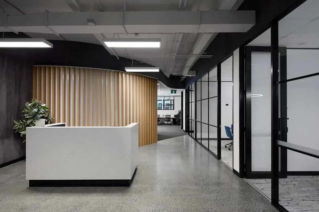 Modern office reception area with a white desk, potted plant, wood slat wall, and glass-walled conference rooms.