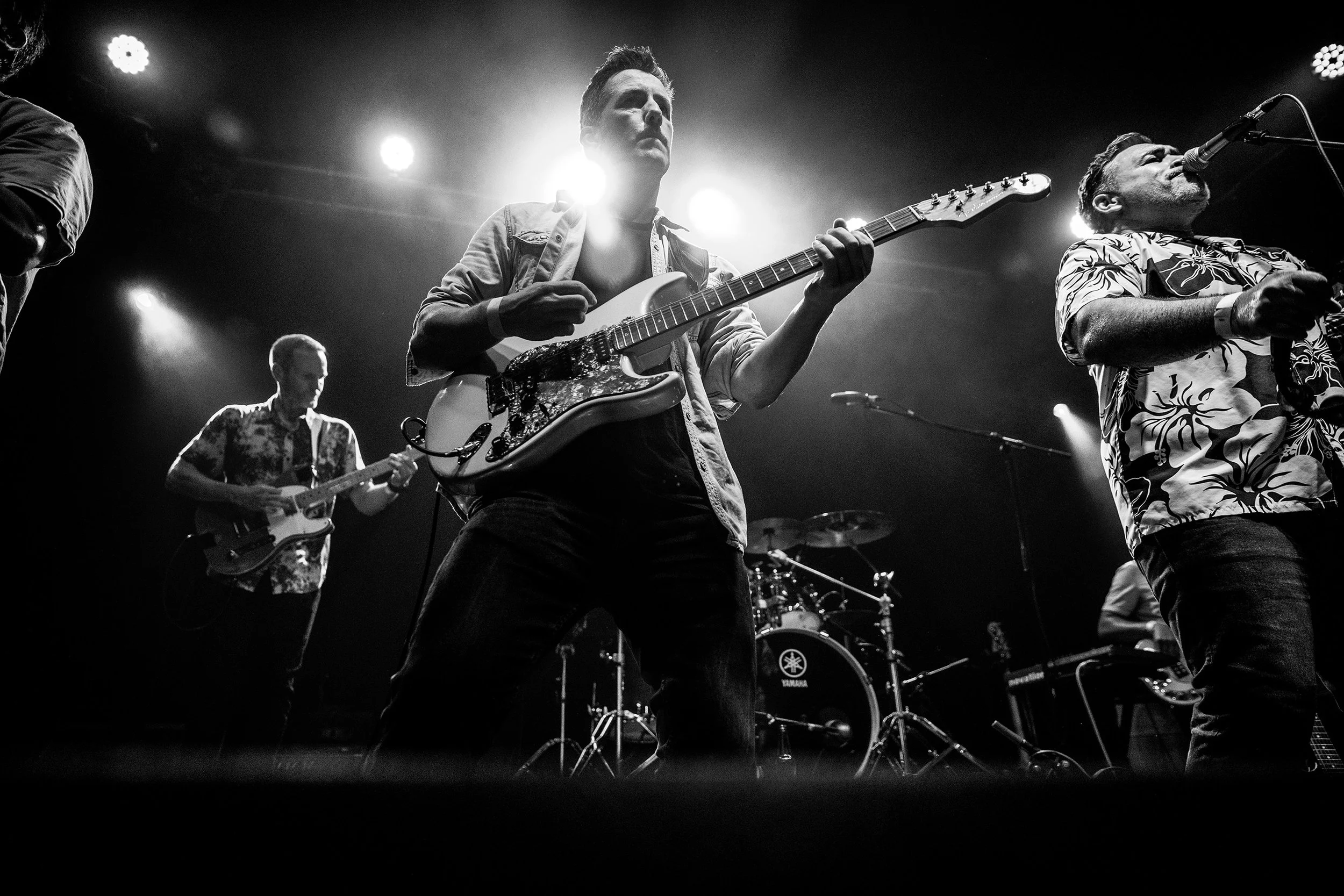 Black and white photo of a band performing on stage with multiple musicians playing guitars and drums, under stage lights.wedding, acoustic musician, wedding DJ, new zealand , tauranga musician, tahuna breaks, wedding music, new zealand wedding music