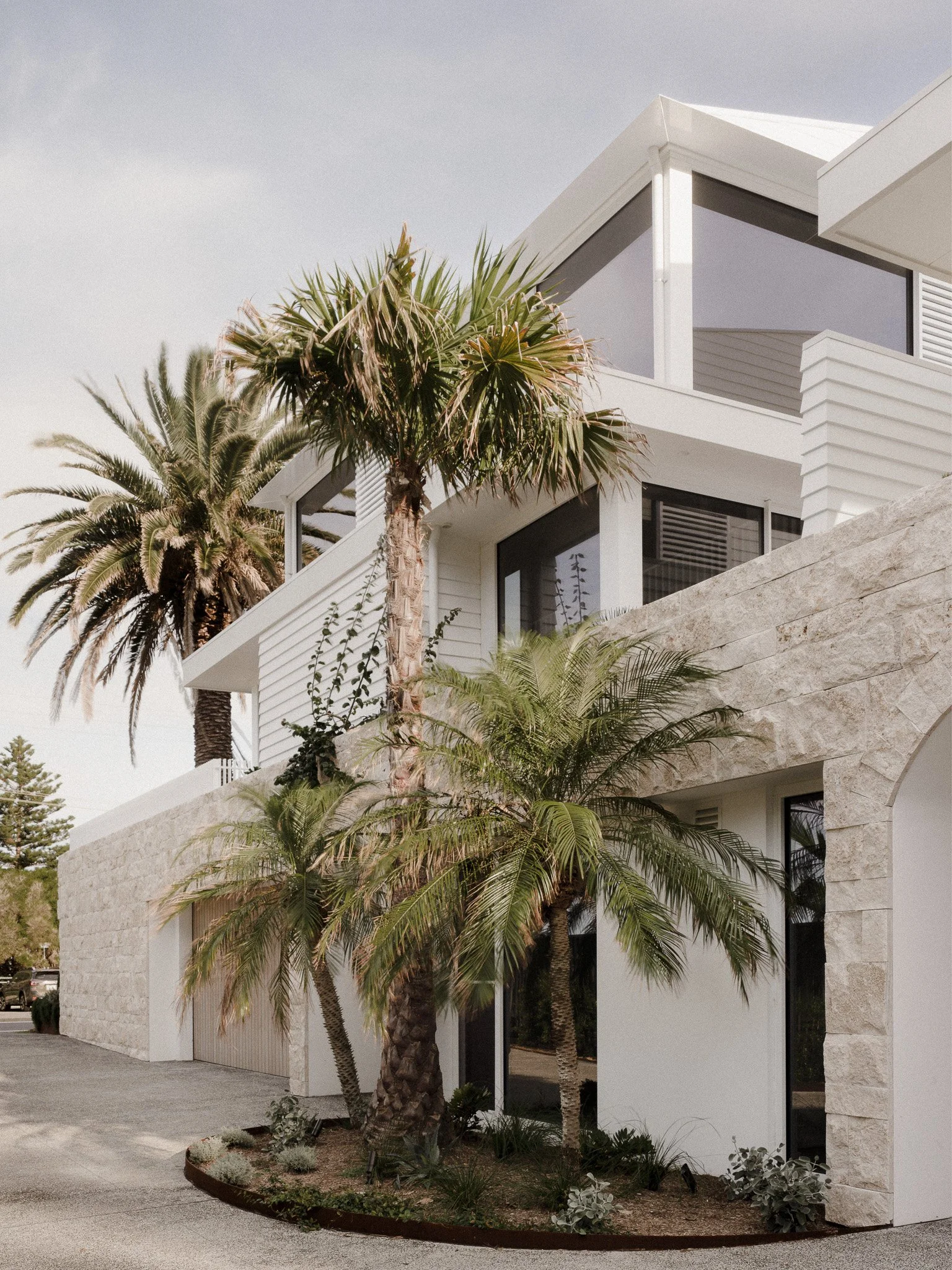 Modern white house with large windows, surrounded by palm trees and desert plants, with a stone wall in the foreground.