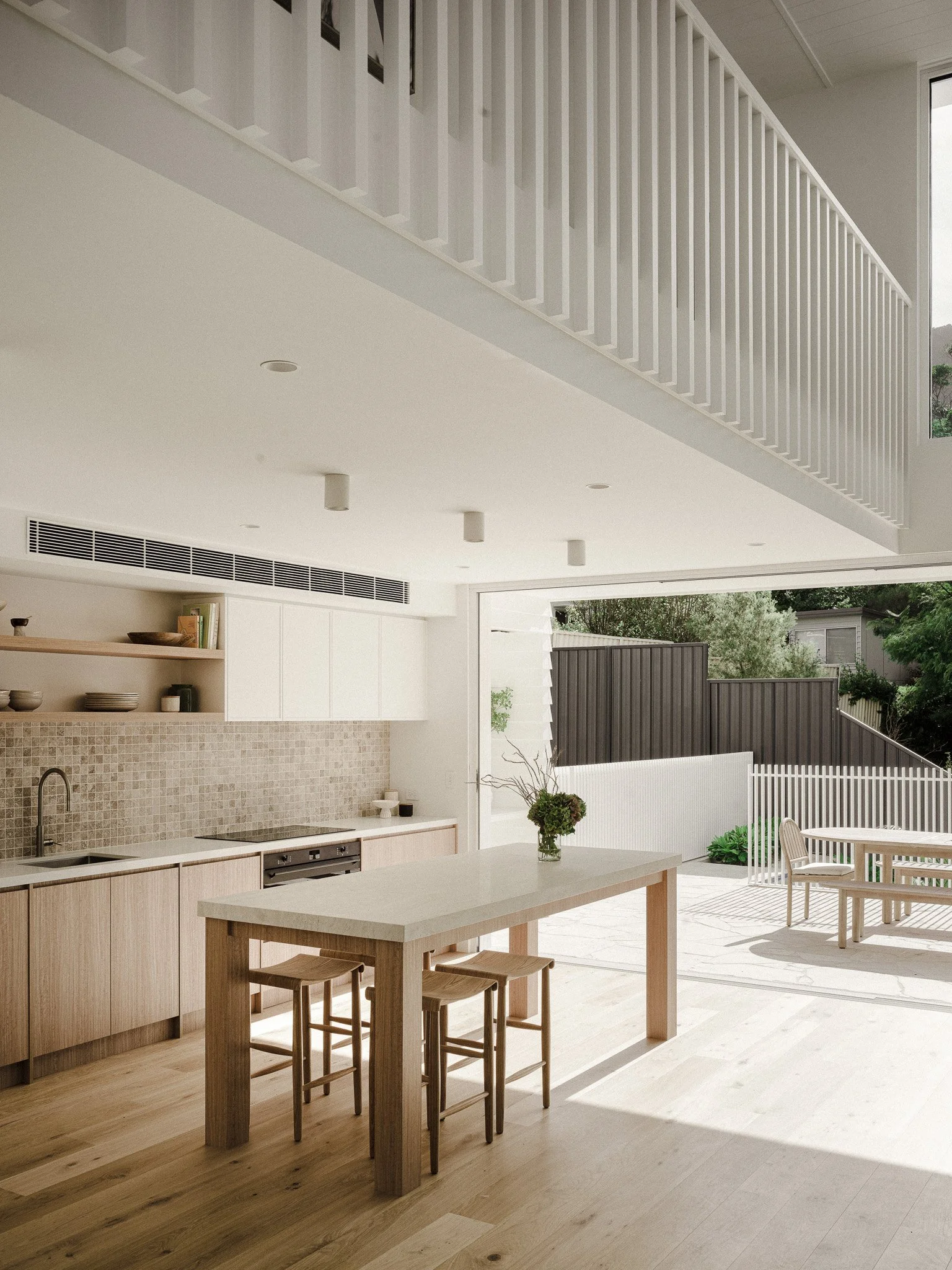 Modern kitchen and dining area with a wooden table and stools, open to an outdoor patio with seating and greenery.
