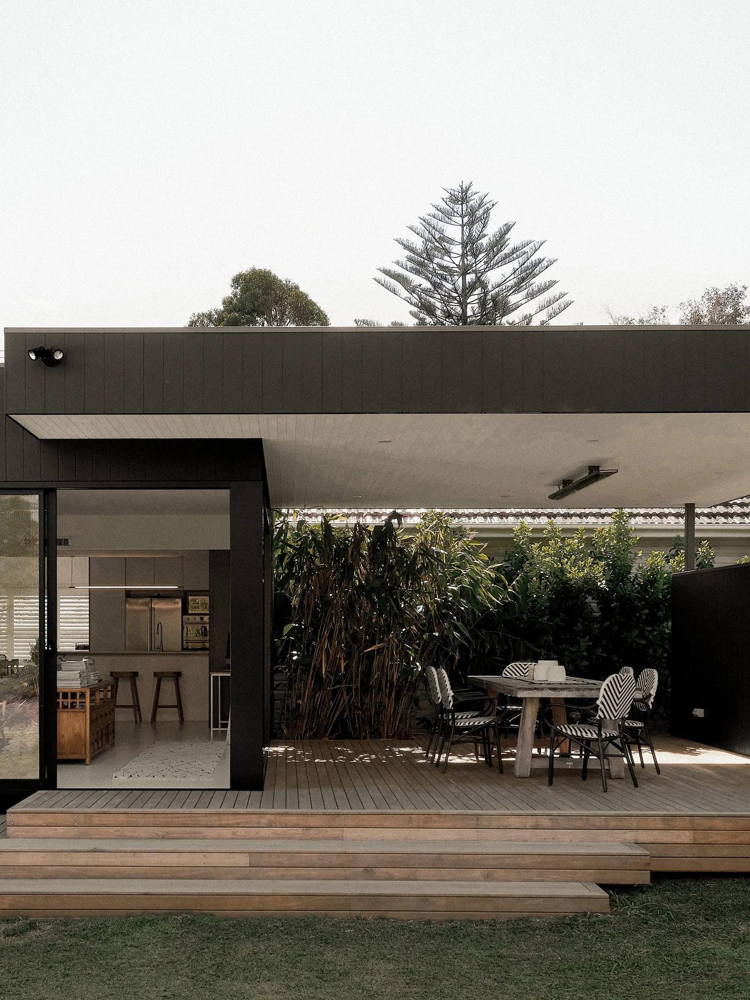 A modern outdoor patio with a dining area, black metal chairs with patterned cushions, a stone table, surrounded by green plants and a wooden deck, with a house and trees in the background under an overcast sky.