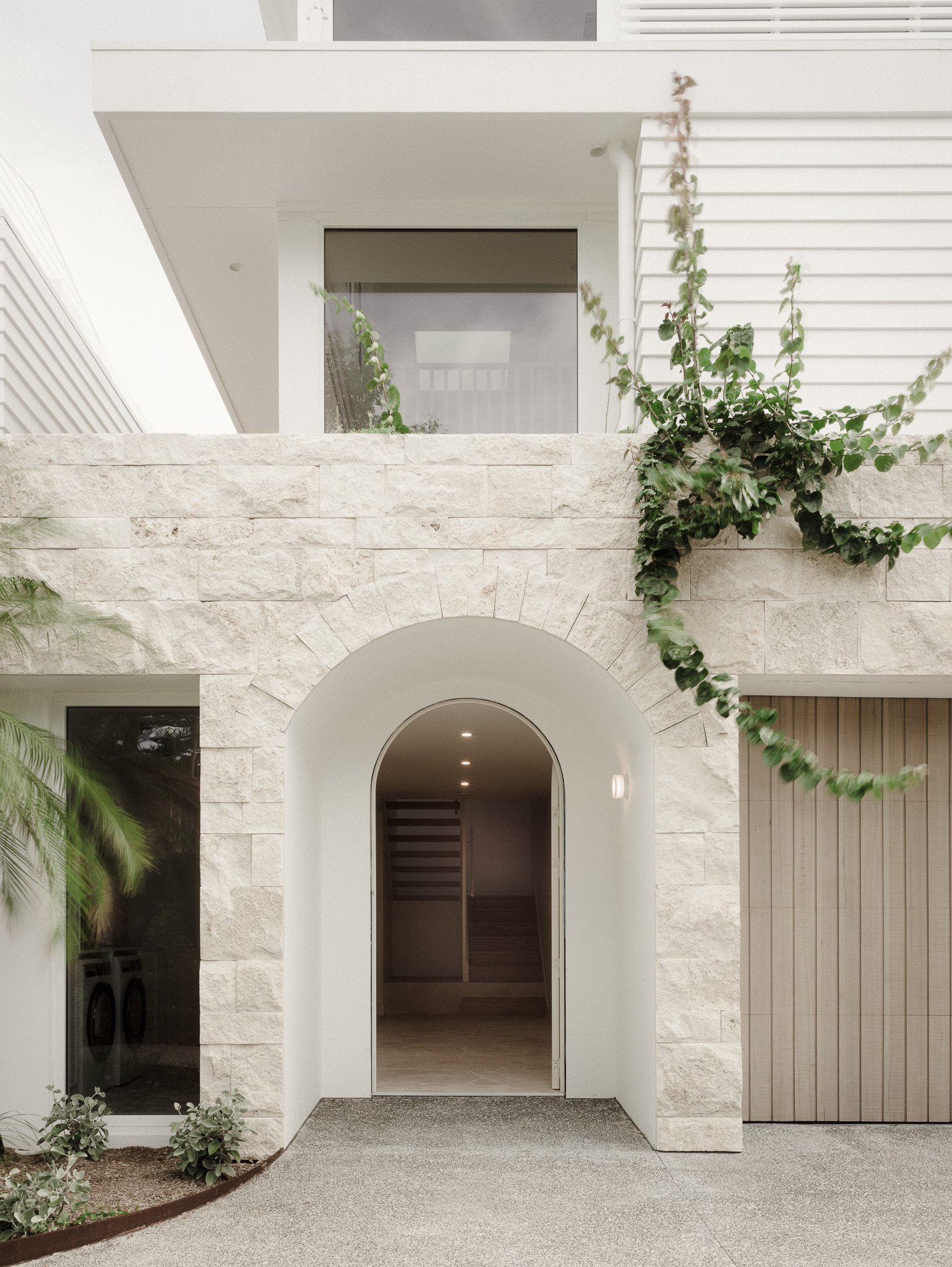 Modern house exterior with a stone archway entrance, white walls, a glass door, and a balcony with greenery.