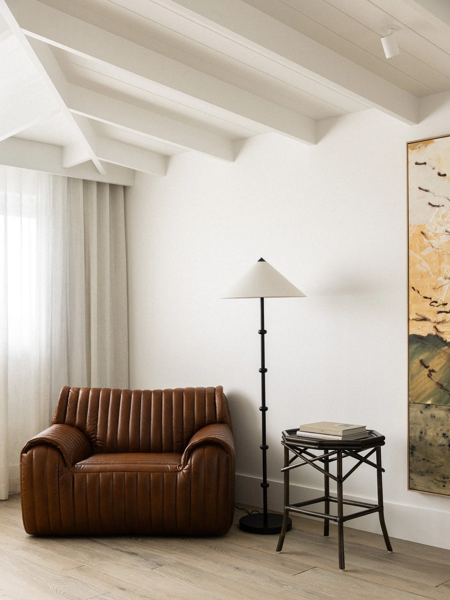 A living room corner with a leather armchair, a floor lamp, a side table with books, a window with curtains, a wall with art, and wooden flooring.