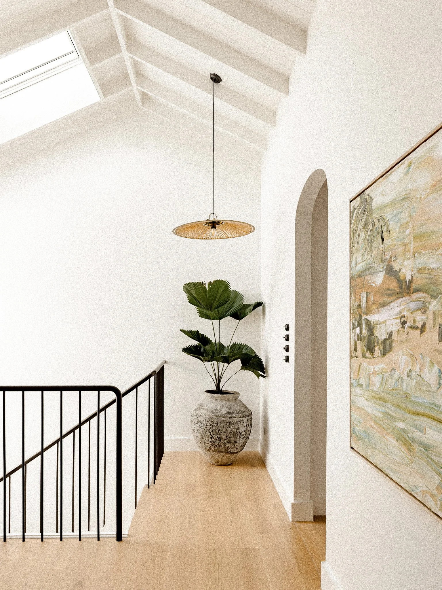 A modern indoor space with white walls and a beige floor. There is a large potted plant in a textured gray vase, a hanging pendant light with a wide, woven shade, a black metal staircase railing, a colorful abstract painting on the right, and an arch