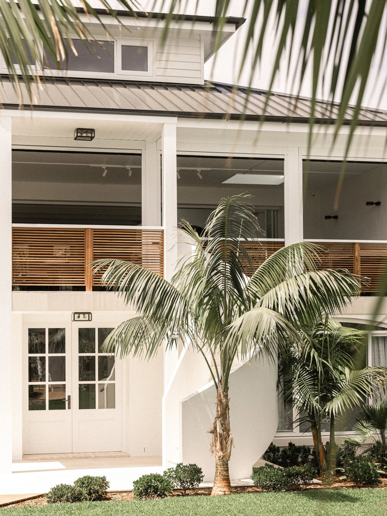 A modern residential house facade with a white exterior, a glass-paned front door, large windows, a balcony with wooden railing, a palm tree in front, and landscaped bushes and grass.