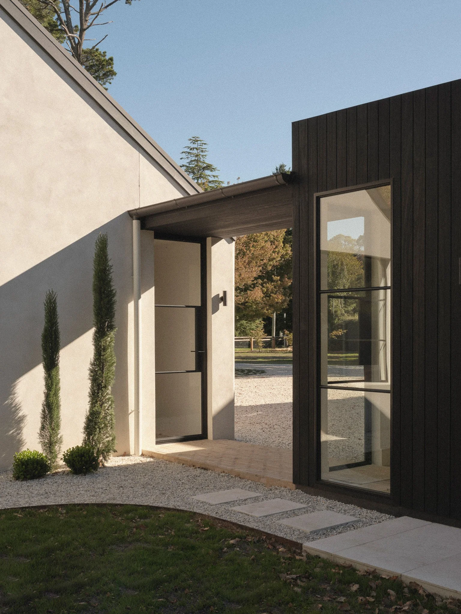 Modern house exterior with black and white walls, large glass windows, and landscaped yard with grass, small bushes, and a pathway of large stone slabs.