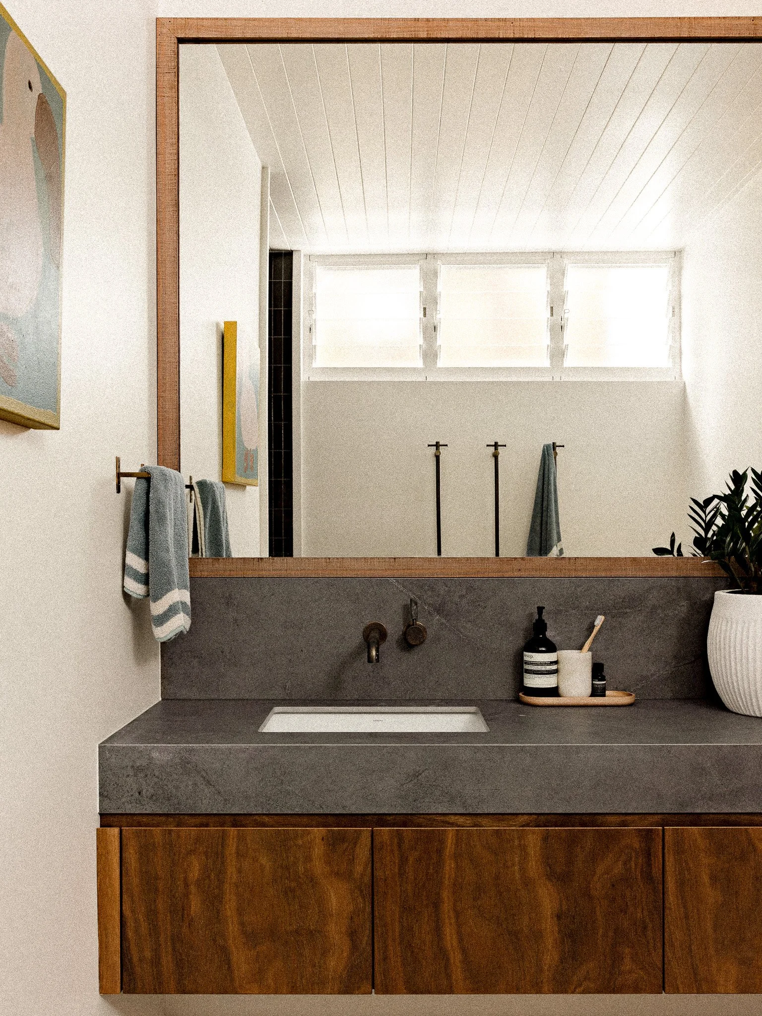 A modern bathroom vanity with a rectangular white sink installed in a dark gray stone countertop, above a wooden cabinet with a warm finish. On the countertop, there are black soap and lotion bottles, a toothbrush, and a small tray. A large mirror wi