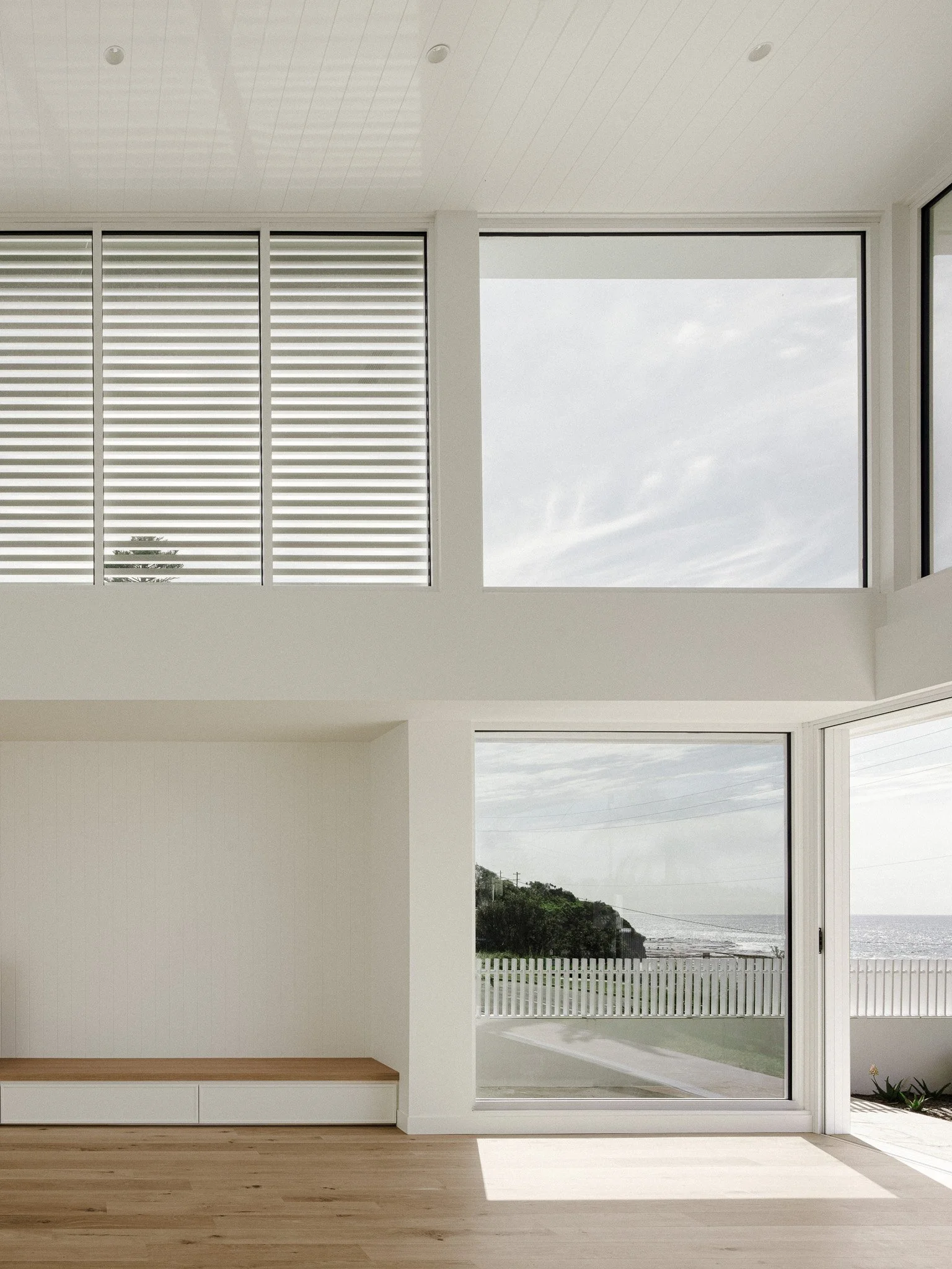 Empty living room with large windows overlooking a coastline with trees and fencing.