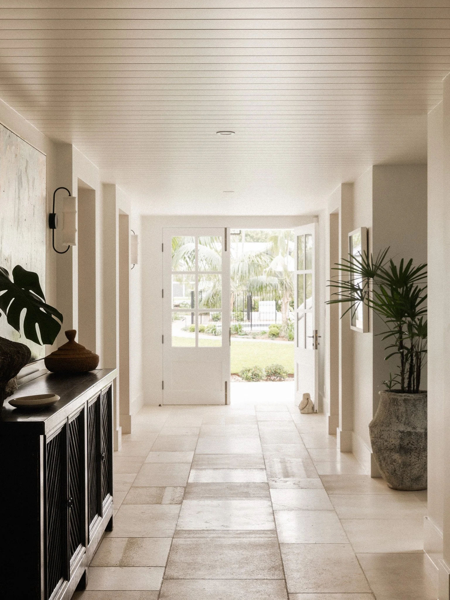 Bright hallway with large window and door at the end, decorated with plants and wall art, beige tiled floor, and white walls and ceiling.