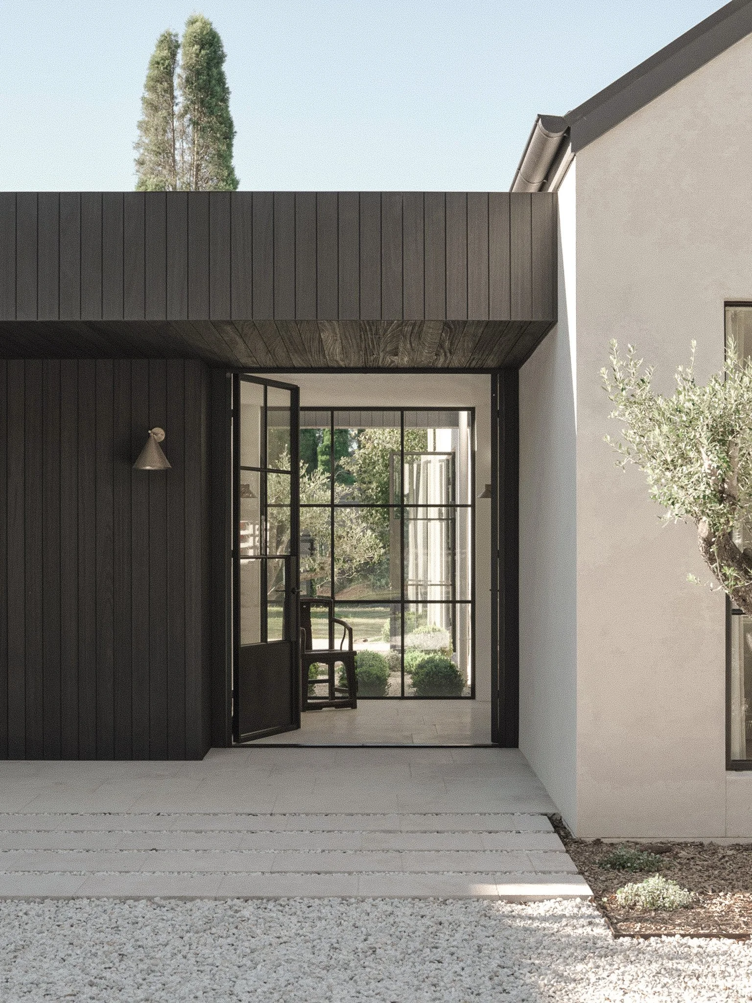 View through black-framed glass doors into an interior space with a chair and a large window showing outdoor greenery with trees and shrubs. The exterior wall is white, and part of a small garden with gravel, plants, and a tree is visible in the fore