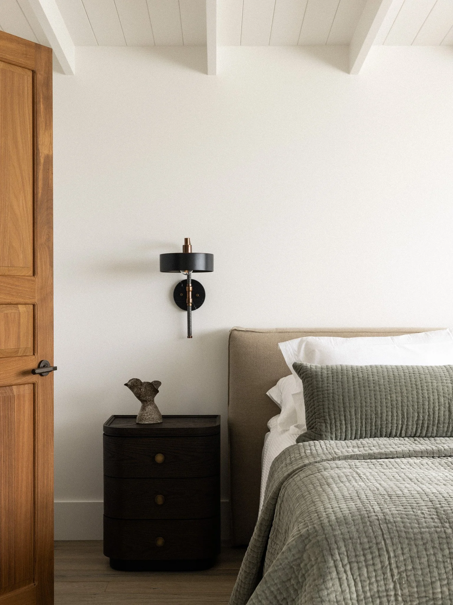 A bedroom with a bed with gray quilted bedding and a beige headboard, a black nightstand with a decorative sculpture, a black wall-mounted lamp, a white wall, and wooden flooring.