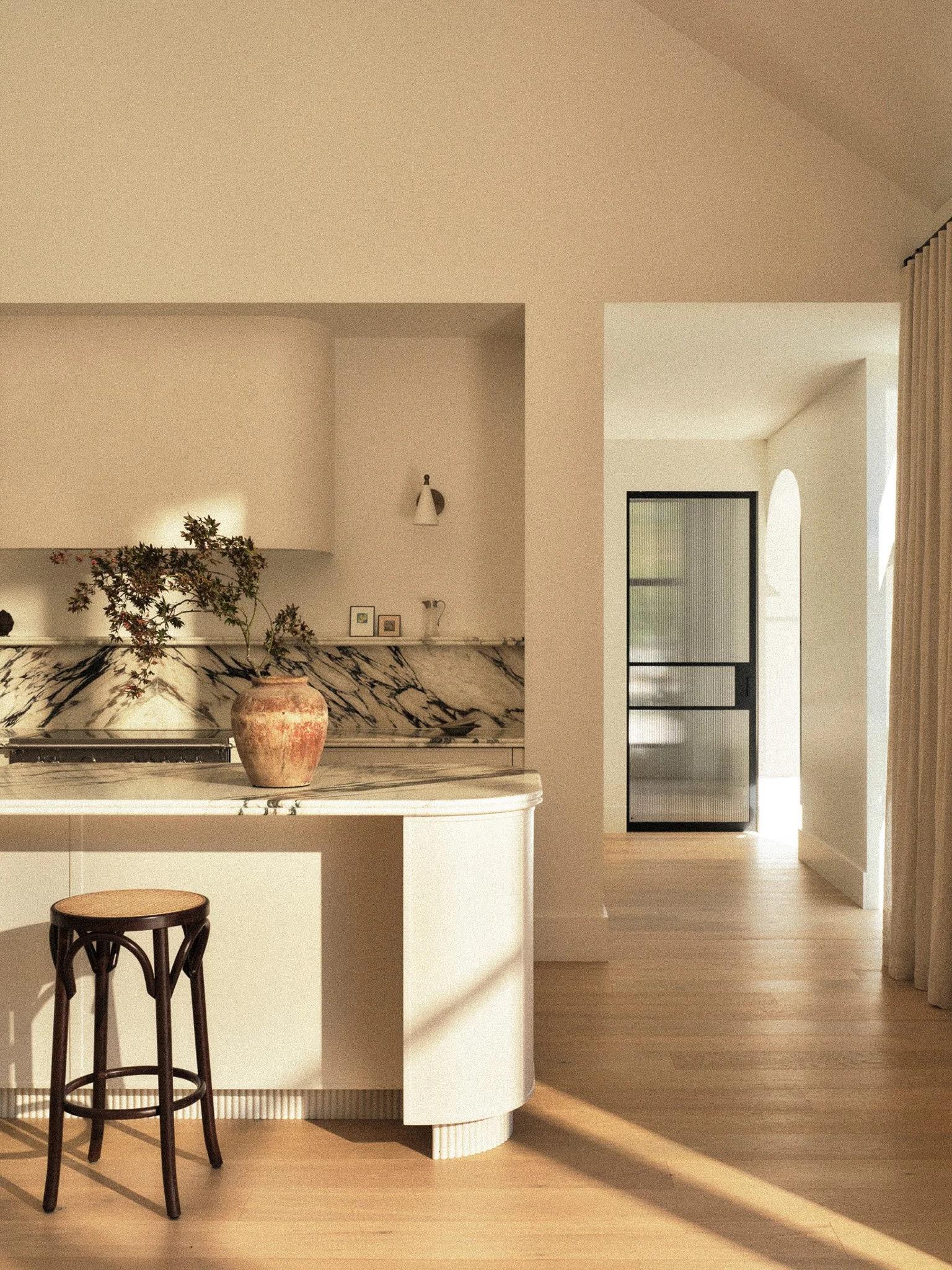 Interior view of a modern kitchen with a marble island, two dark wooden bar stools, a large ceramic vase with a plant, and a doorway leading to another room with a large window and curtains, in warm natural lighting.