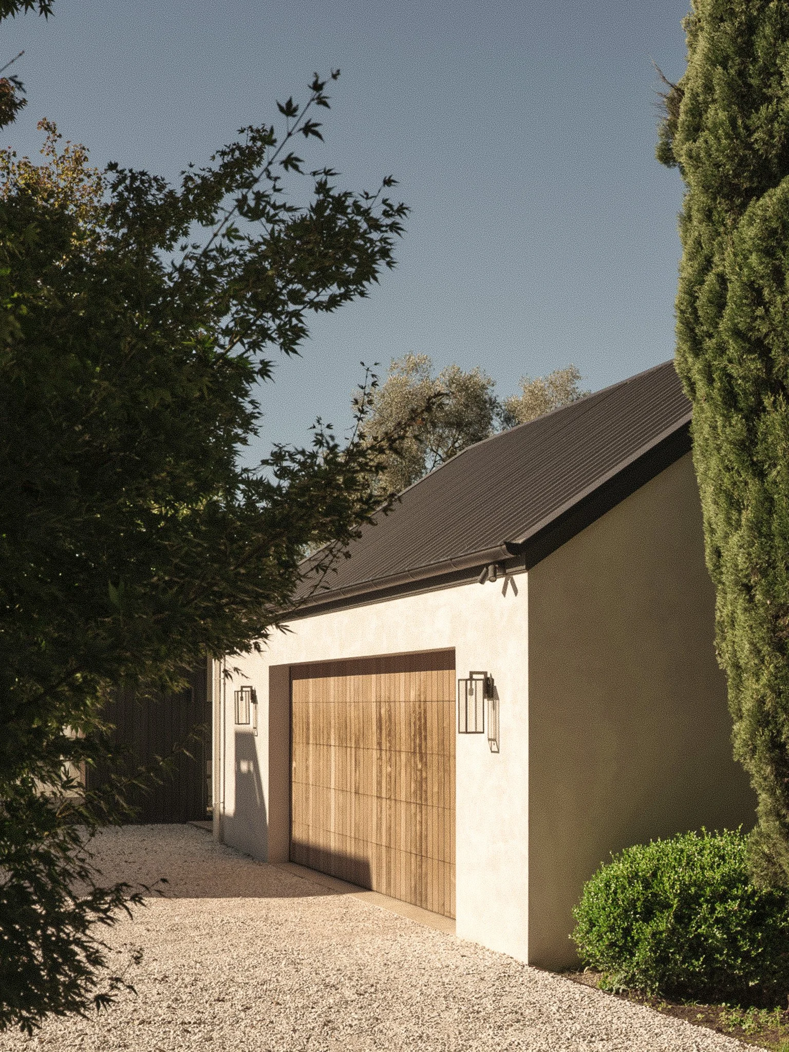Modern garage with a large wooden door, surrounded by trees and bushes, with a gravel driveway.