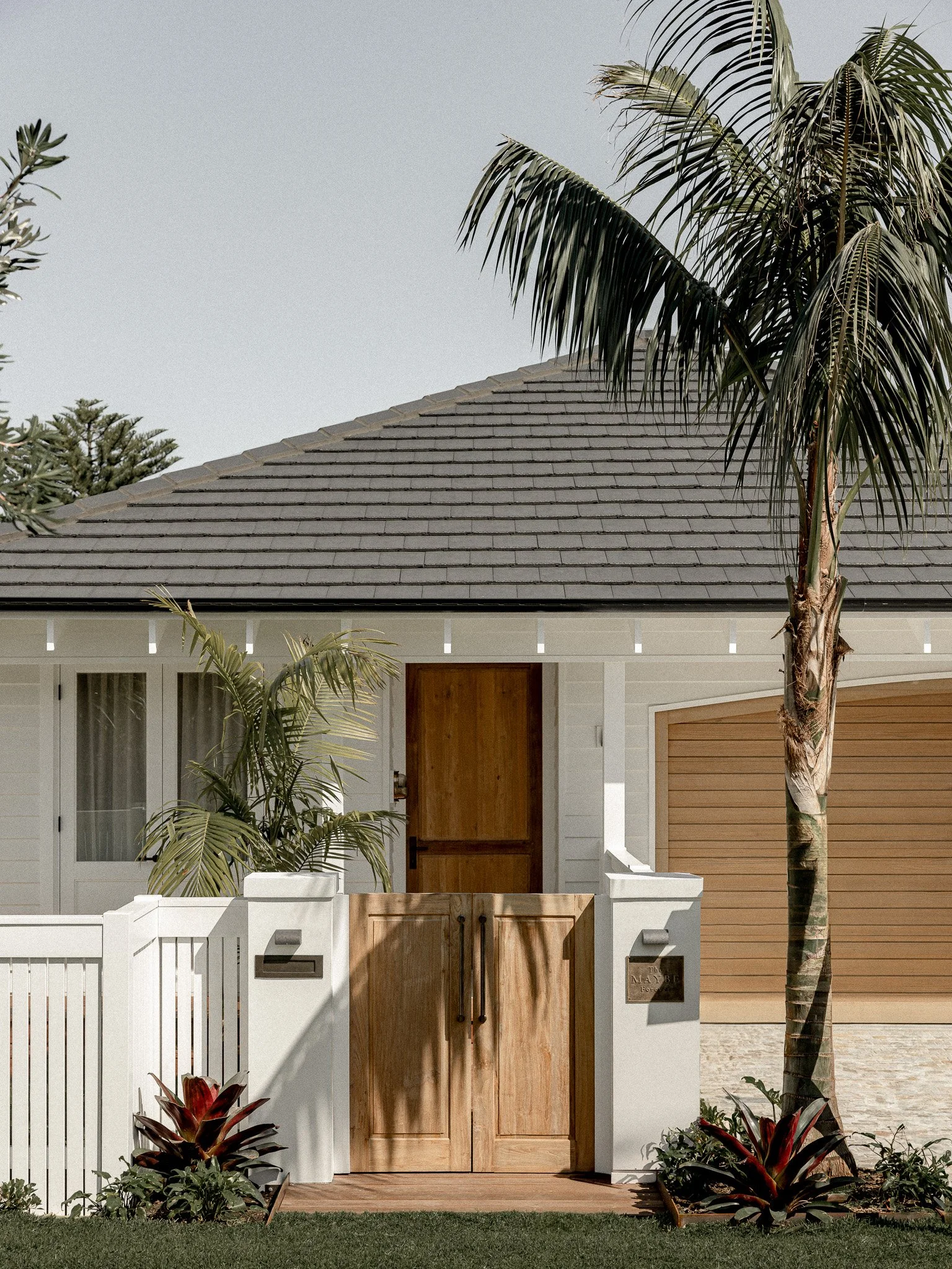 Front view of a modern house facade with a gabled roof, wooden door, white exterior walls, a small front garden with tropical plants and a tall palm tree, and a wooden garage door on the right.