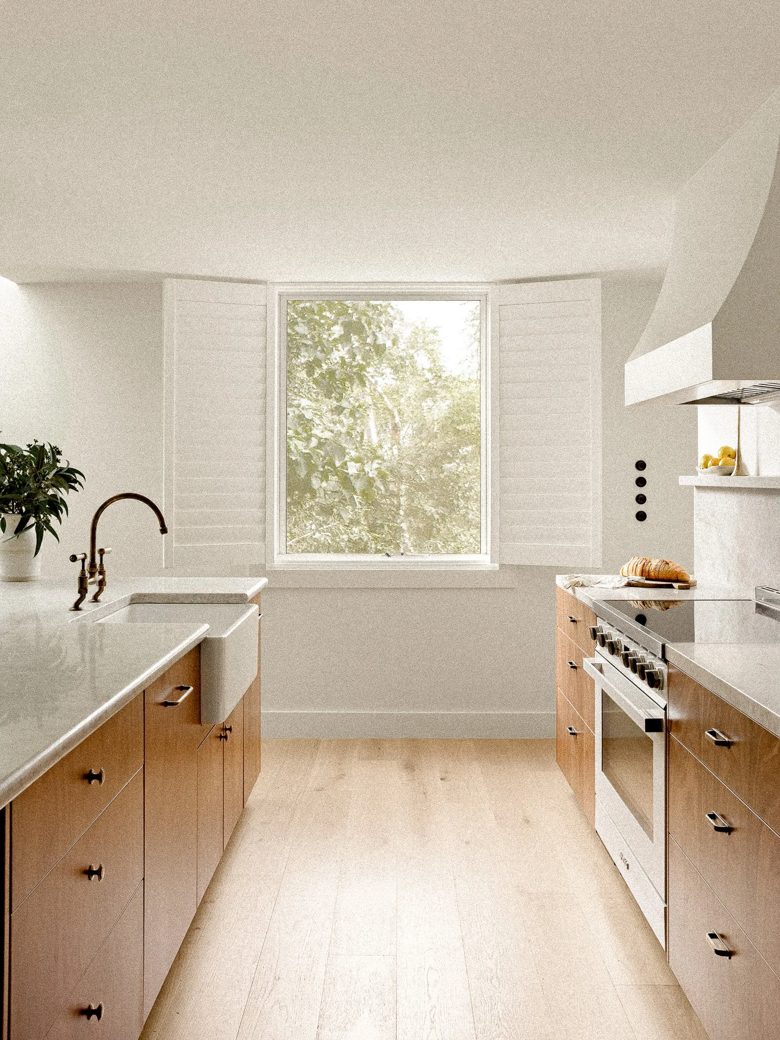 A modern, minimalistic kitchen with wooden cabinets, a white countertop, a stove, a window with green trees outside, and a potted plant on the counter.