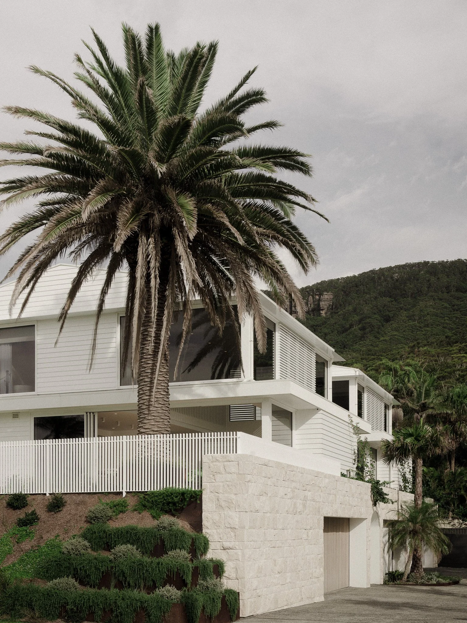 A modern white multi-story house with large windows and a flat roof, surrounded by a white picket fence, a large palm tree in the front yard, landscaped greenery, and a mountainous background with trees under a cloudy sky.