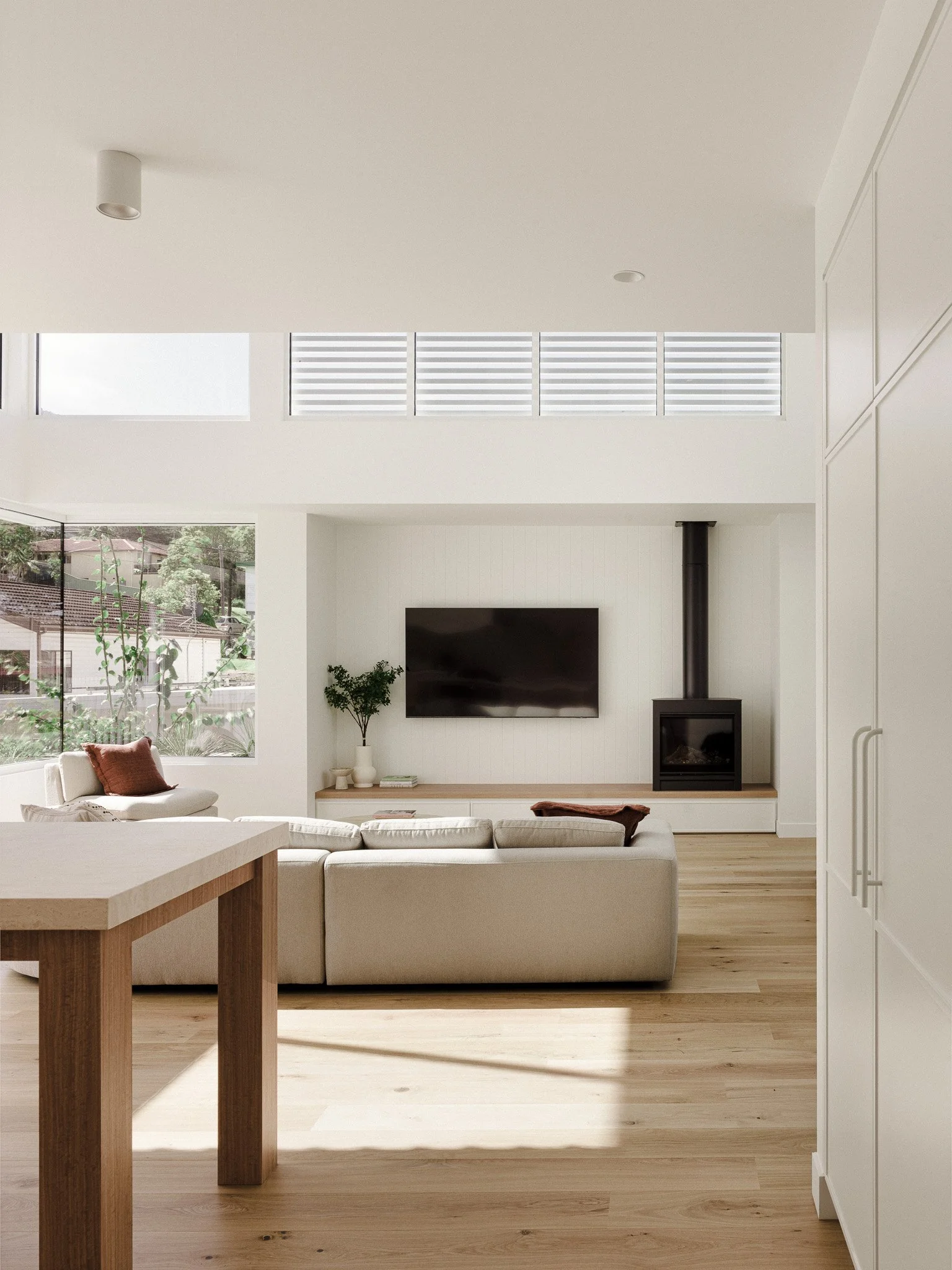 Modern living room with large window, white sofa, wall-mounted TV, wood stove, and minimal decor.