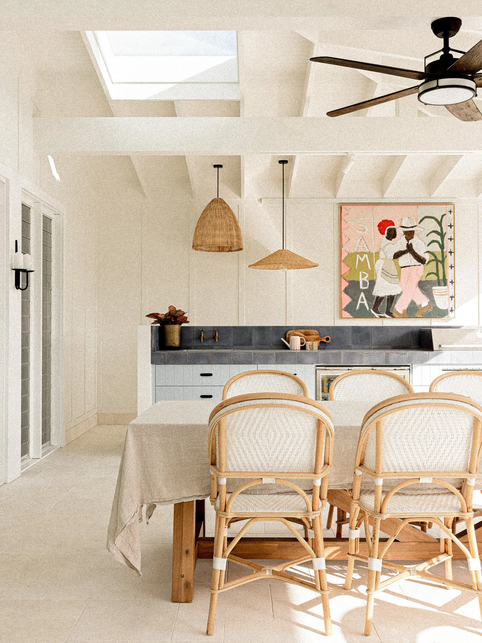 Bright kitchen with white walls, cream tile flooring, a wooden dining table with cream upholstered chairs, a skylight, hanging wicker lights, and wall art depicting two women in traditional clothing.