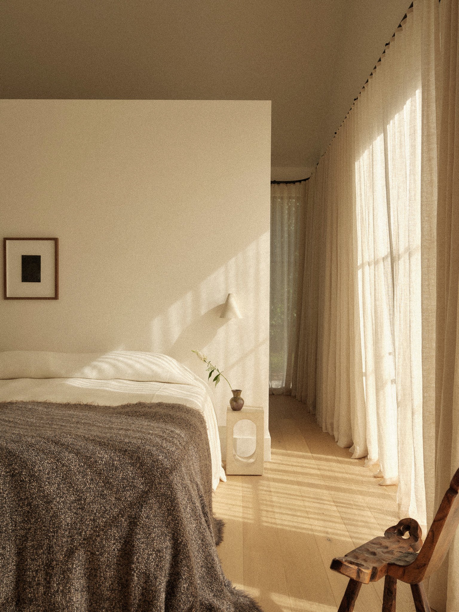 Cozy bedroom with a bed covered in a dark textured blanket, a small white nightstand with a decorative vase, sheer curtains allowing sunlight to fill the room, and a rustic wooden chair in the corner.