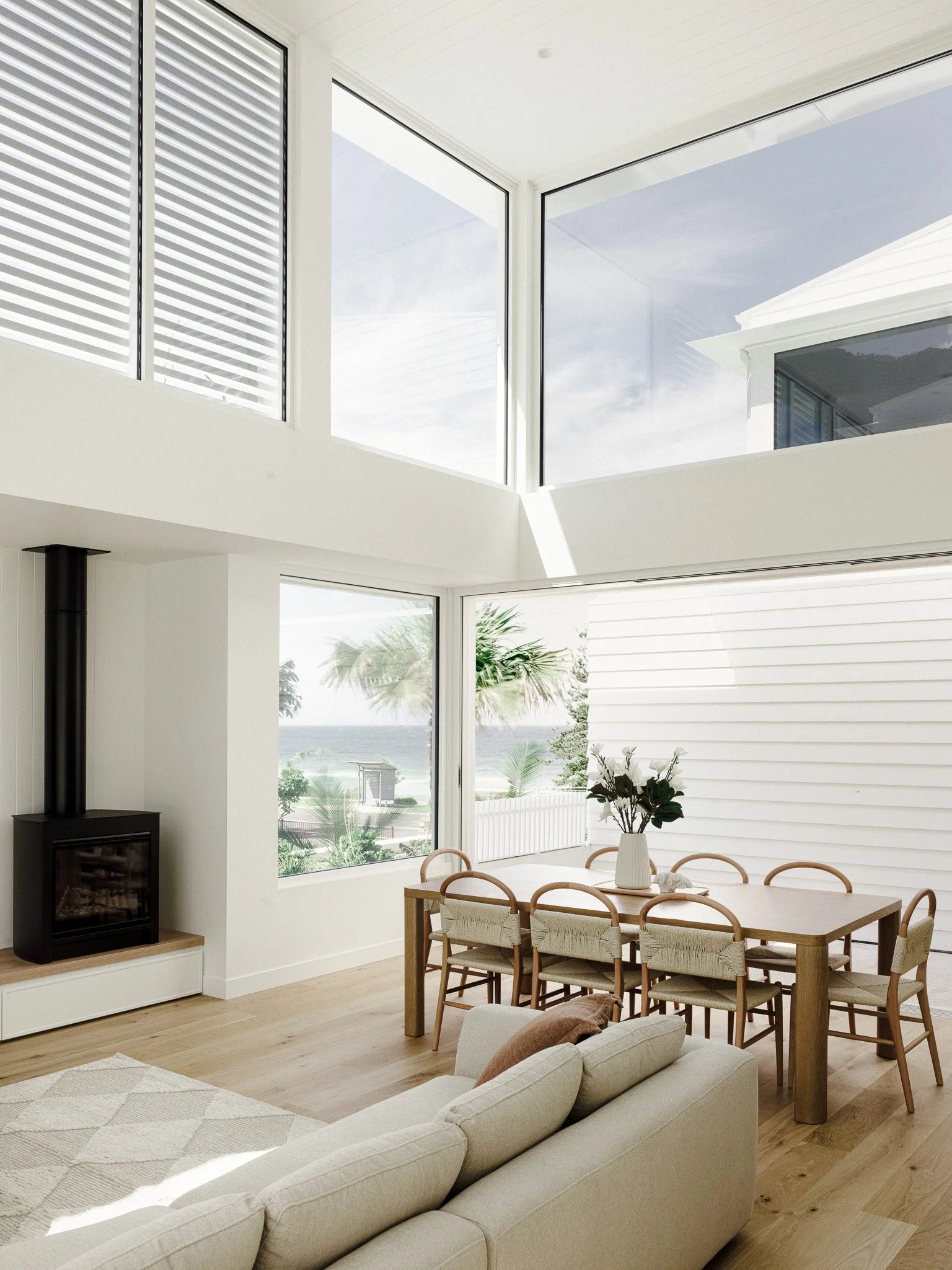 A bright, modern living room with large windows showing a view of the beach and ocean. The room has a beige sofa, a wooden dining table with chairs, a white vase with plants on the table, and a black wood stove. Light-colored wooden flooring and whit