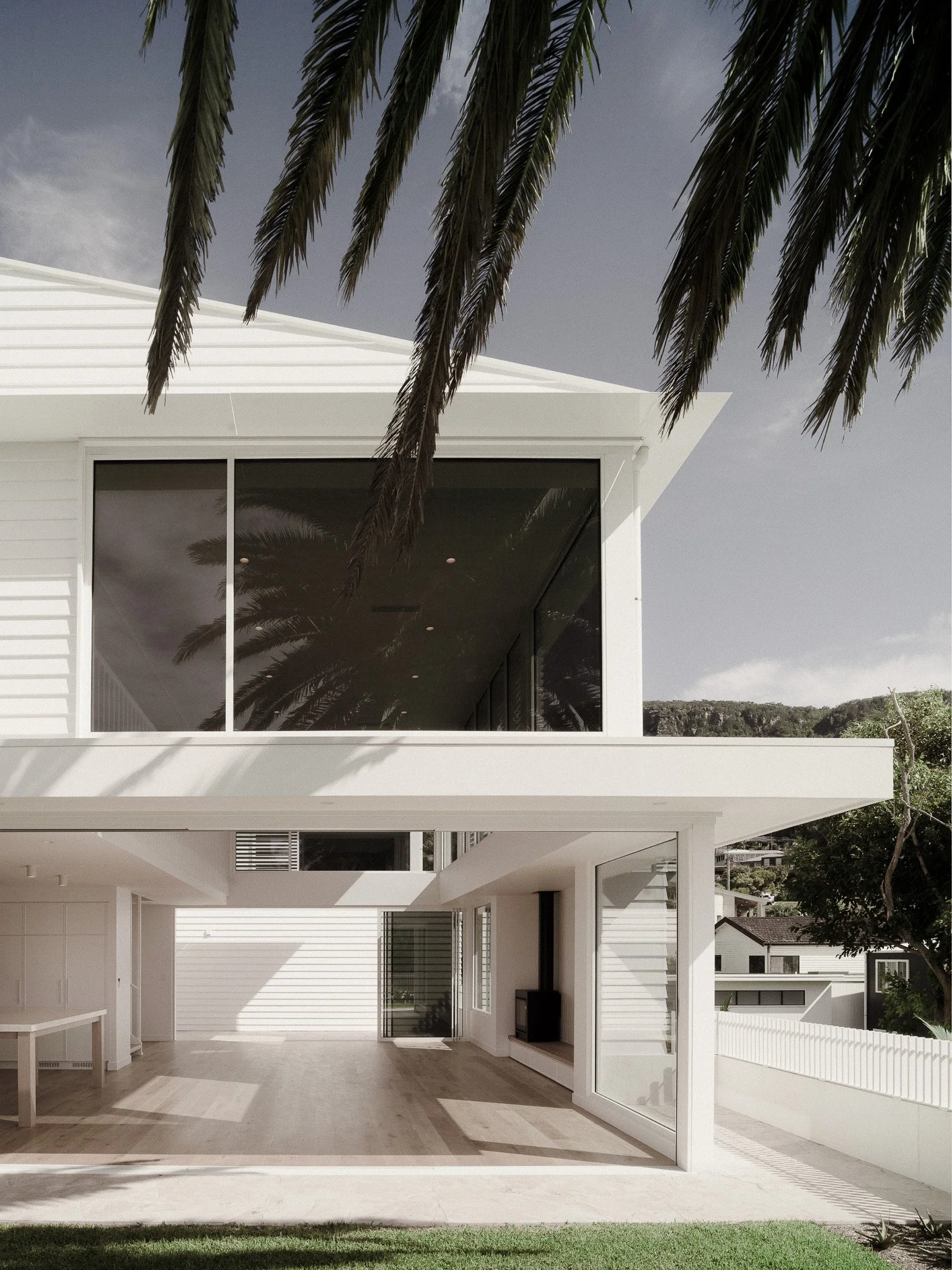 Modern white two-story house with large glass windows and a backyard with green grass. Palm tree branches are visible at the top of the image, set against a partly cloudy sky.