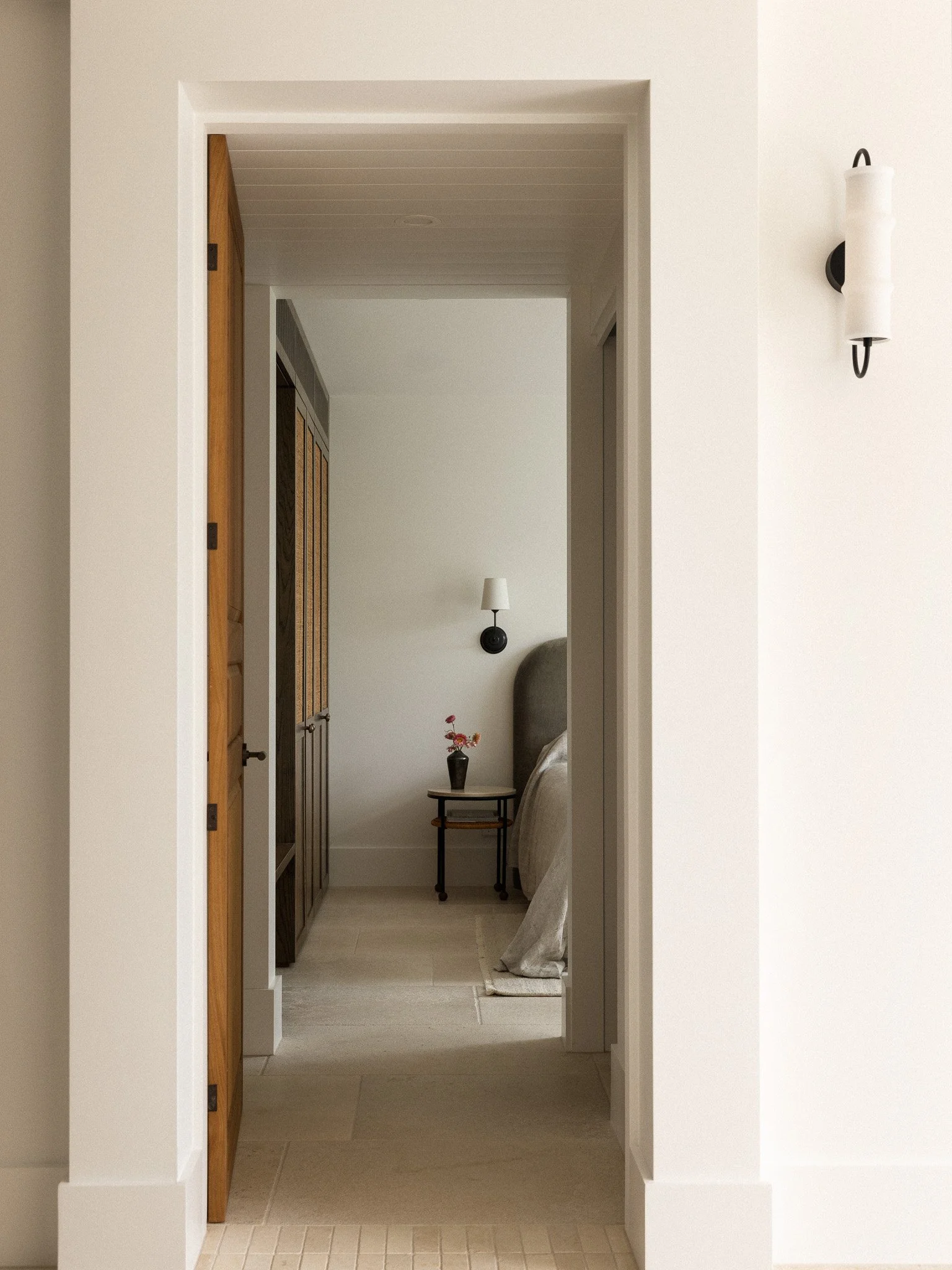 Interior view of a doorway leading to a small room with a side table, a vase with flowers, a standing lamp, and a bed with bedding.