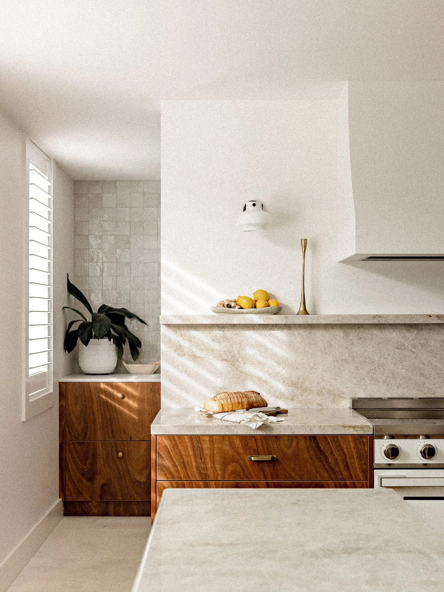 A kitchen counter with a loaf of bread on a paper towel, a cabinet with a potted plant on top, and a bowl of lemons on a window sill in a modern, minimalist kitchen with white walls and wood accents.
