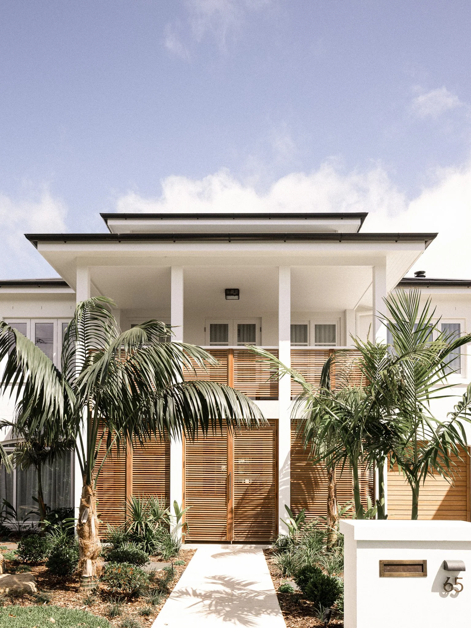 Modern white house with wooden slat front gate, surrounded by palm trees and small shrubs, pathway leading up to entrance, house number 465 attached to white wall, cloudy sky overhead. Bahamas style