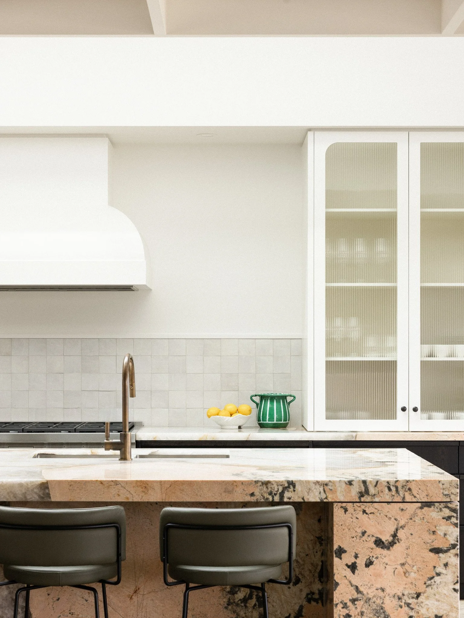 Modern kitchen with white cabinetry, a marble countertop, a stainless steel faucet, a bowl of lemons, and a green pitcher. The backsplash is white tile, and there are dining chairs in the foreground.