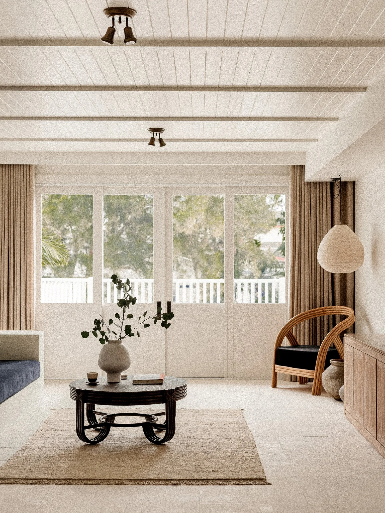 A minimalist living room with light-colored walls and flooring. There is a small round table with a large white vase containing leafy plants. Large windows with beige curtains let in natural light, and a wicker chair with a black cushion is next to a
