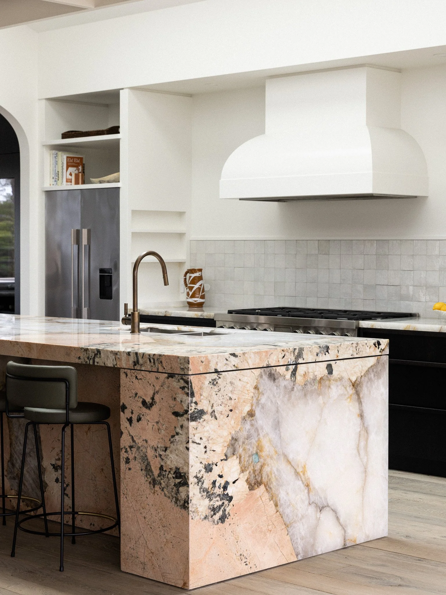 Close-up view of a kitchen island with a beige and black marble countertop and a large natural stone base, with two black barstools.