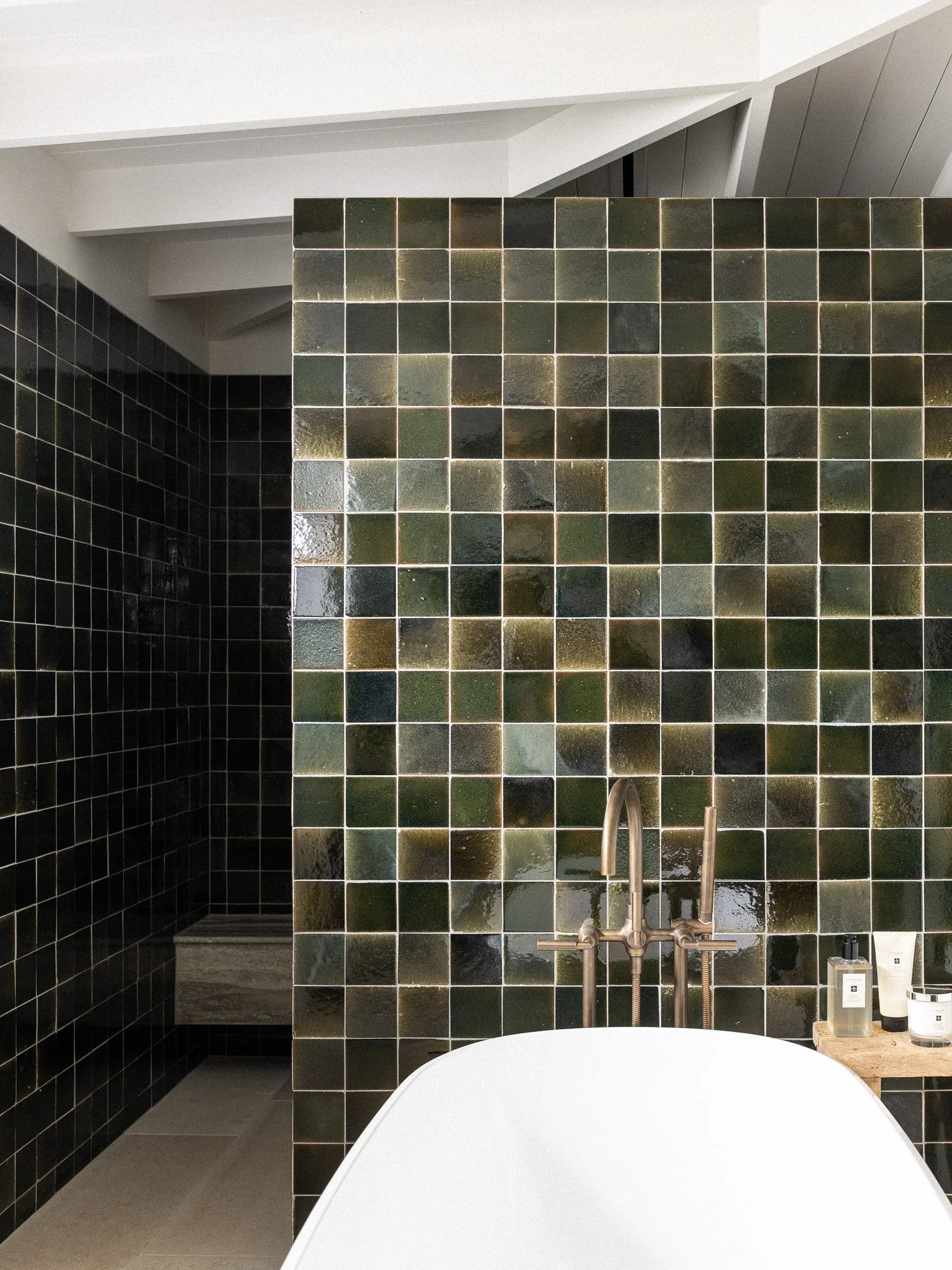 Modern bathroom with black tile walls and a white bathtub in the foreground. A separate shower area with dark green tile walls and a silver shower fixture is visible in the background. The ceiling is white with architectural beams.