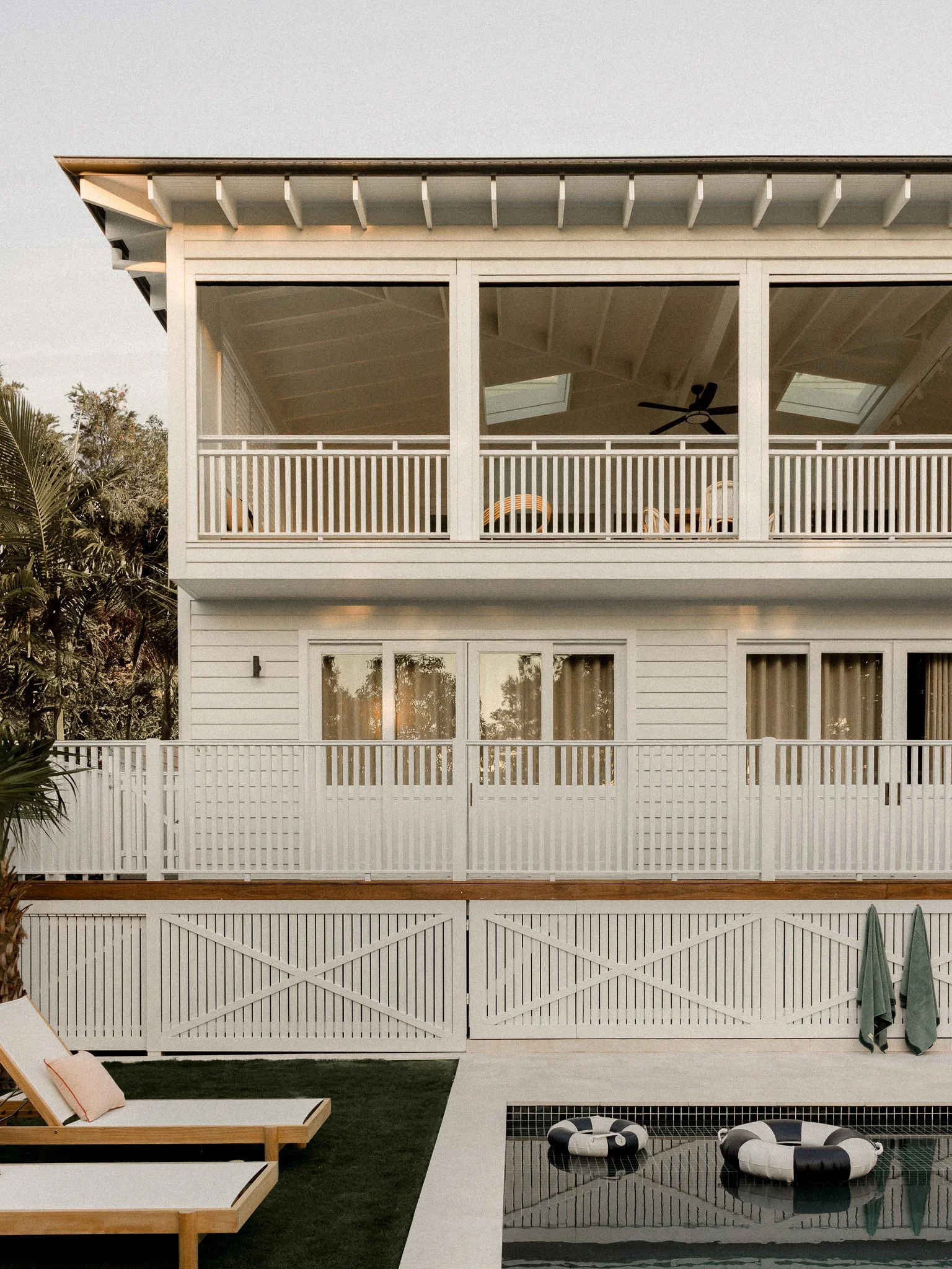 View of a modern house with a balcony, glass doors, a fence, a swimming pool with black and white floats, a lounge chair with a pillow, and trees in the background.
