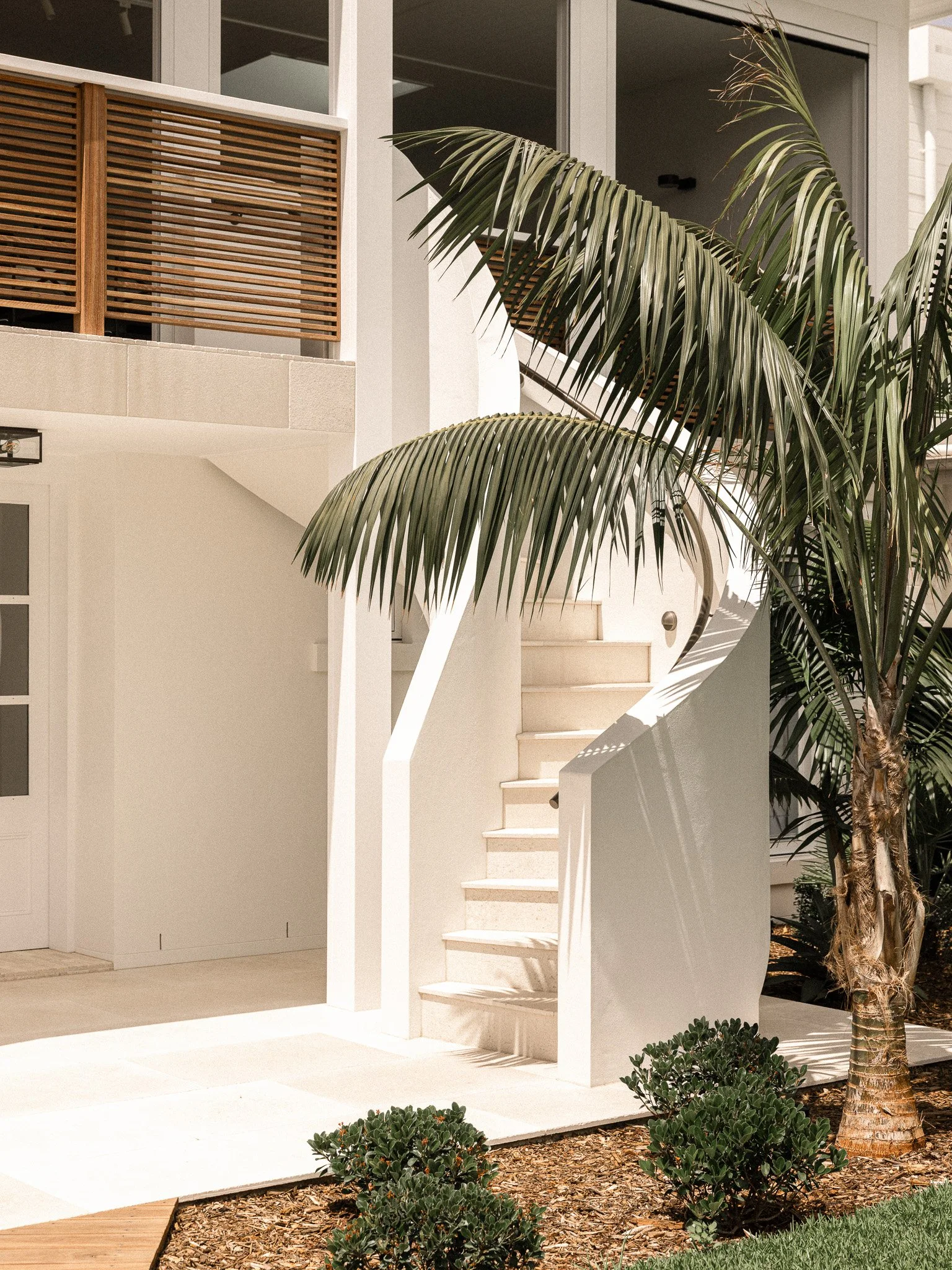 Exterior of a modern white residential building with a staircase, palm trees, and manicured bushes in the front yard.