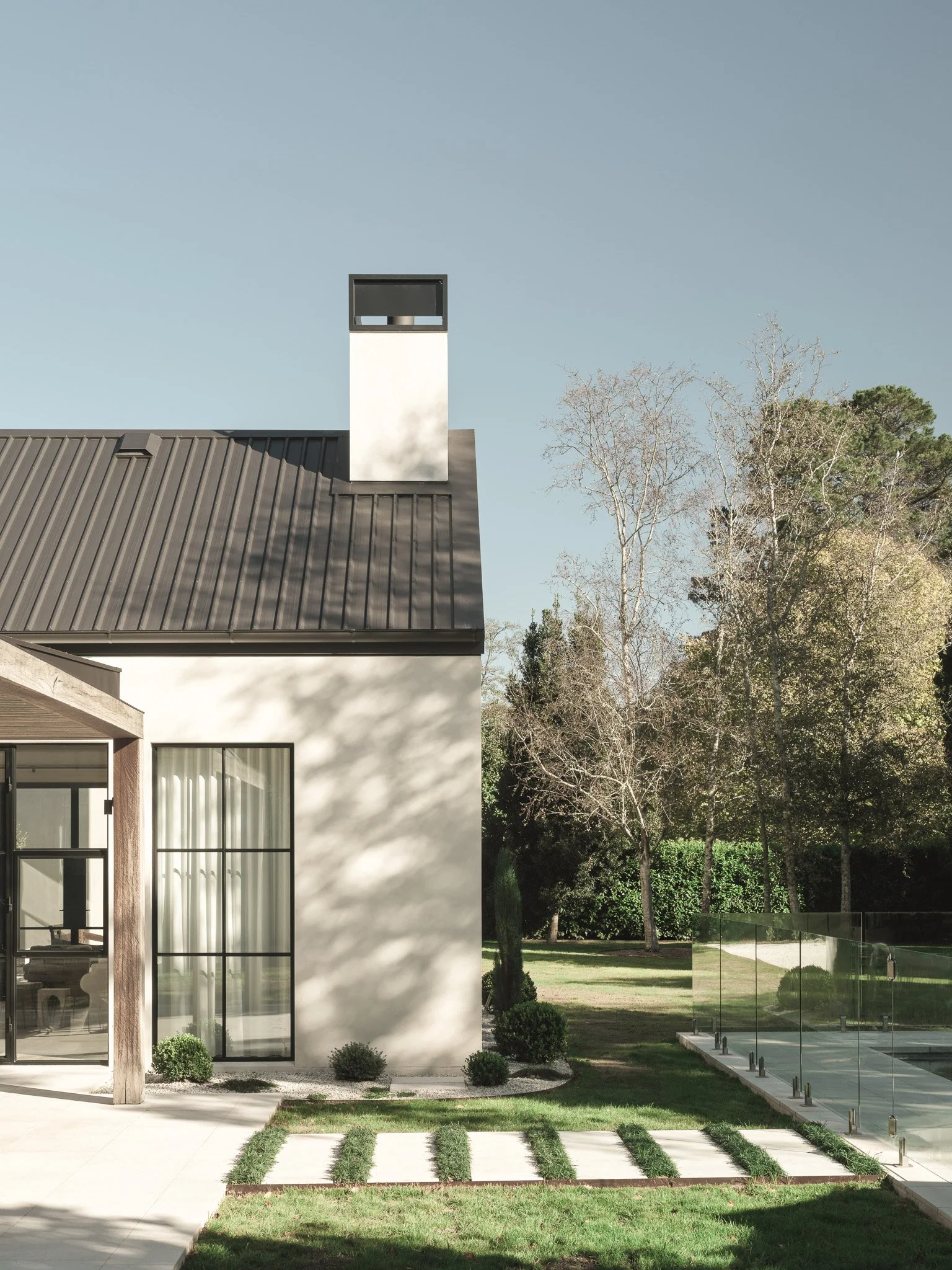 Modern house with large windows, metal roof, and a garden with grass and small bushes, surrounded by trees and a glass fence.