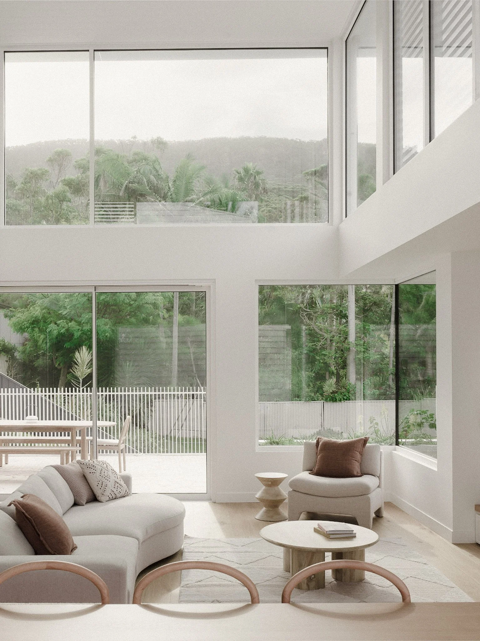 Modern living room with large windows overlooking a lush green landscape, featuring white furniture, throw pillows, and a small round coffee table.