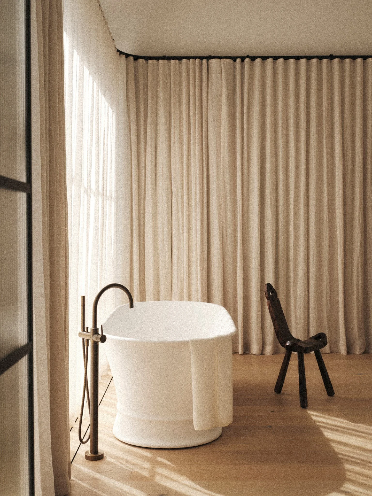 A modern bathroom with a round bathtub, a dark wooden chair, beige curtains, and natural light streaming in.