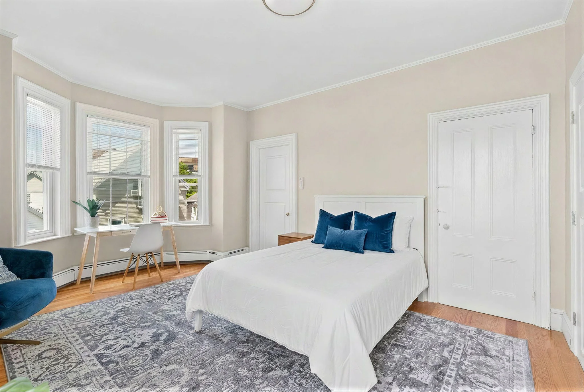 A bright bedroom with a white bed and blue pillows, large windows with blinds letting in natural light, a small desk with a chair and plant near the windows, and a patterned area rug on wooden floor.