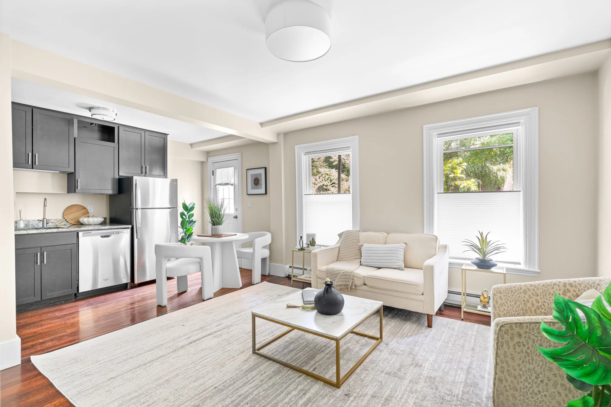 Living room and kitchen area with large windows, beige walls, gray cabinets, stainless steel refrigerator, white dining table with chairs, beige sofa with pillows, coffee table, potted plants, and a white rug on hardwood floor.