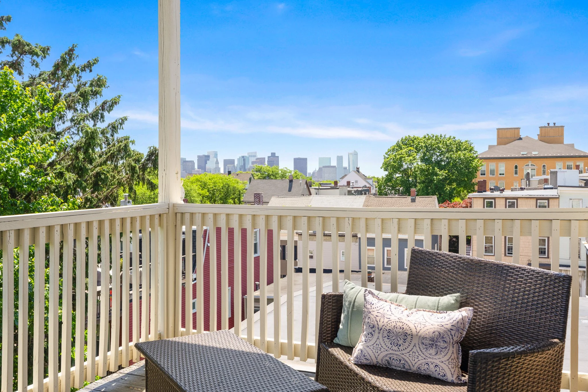 A view from a balcony with wicker chairs and pillows, overlooking rooftops and a city skyline under a clear blue sky with some trees in the foreground.