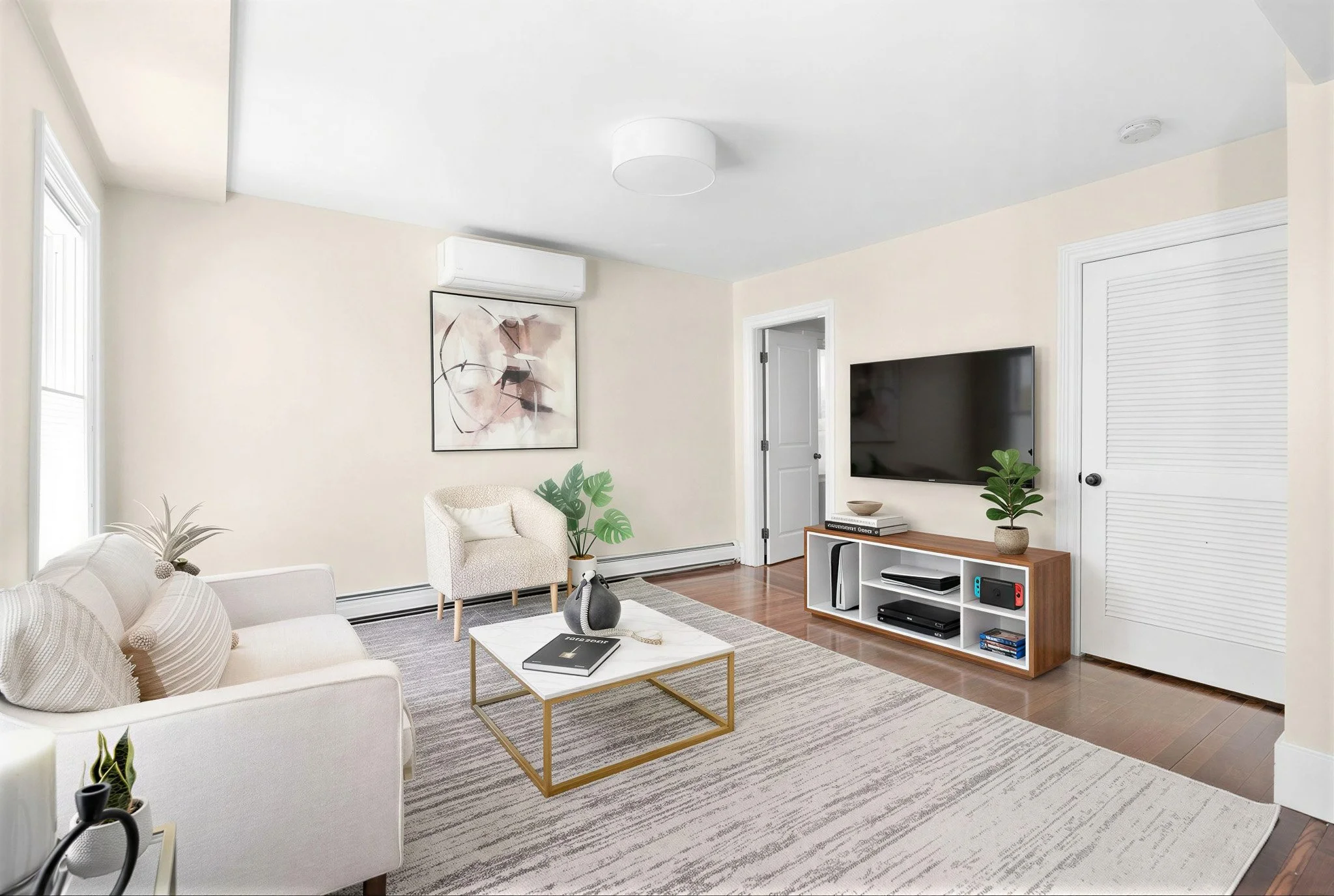 Living room with beige walls, white furniture, a patterned area rug, a wall-mounted flat-screen TV, a small white and wood console table with plants and books, and a window with white blinds.