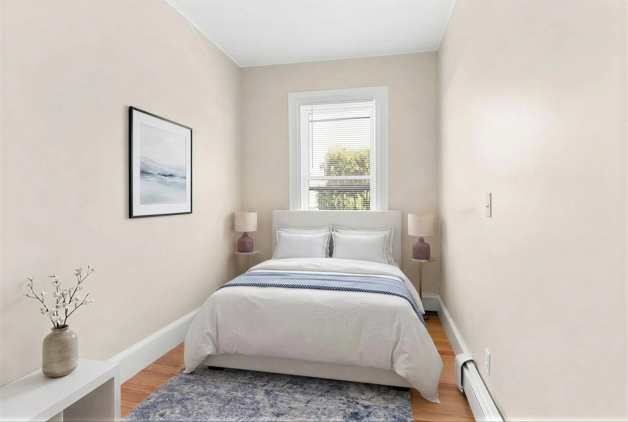 A cozy bedroom with a neatly made bed, two pink bedside lamps, a framed picture on the wall, a window with blinds, a white vase with flowers, and a small rug on a wooden floor.