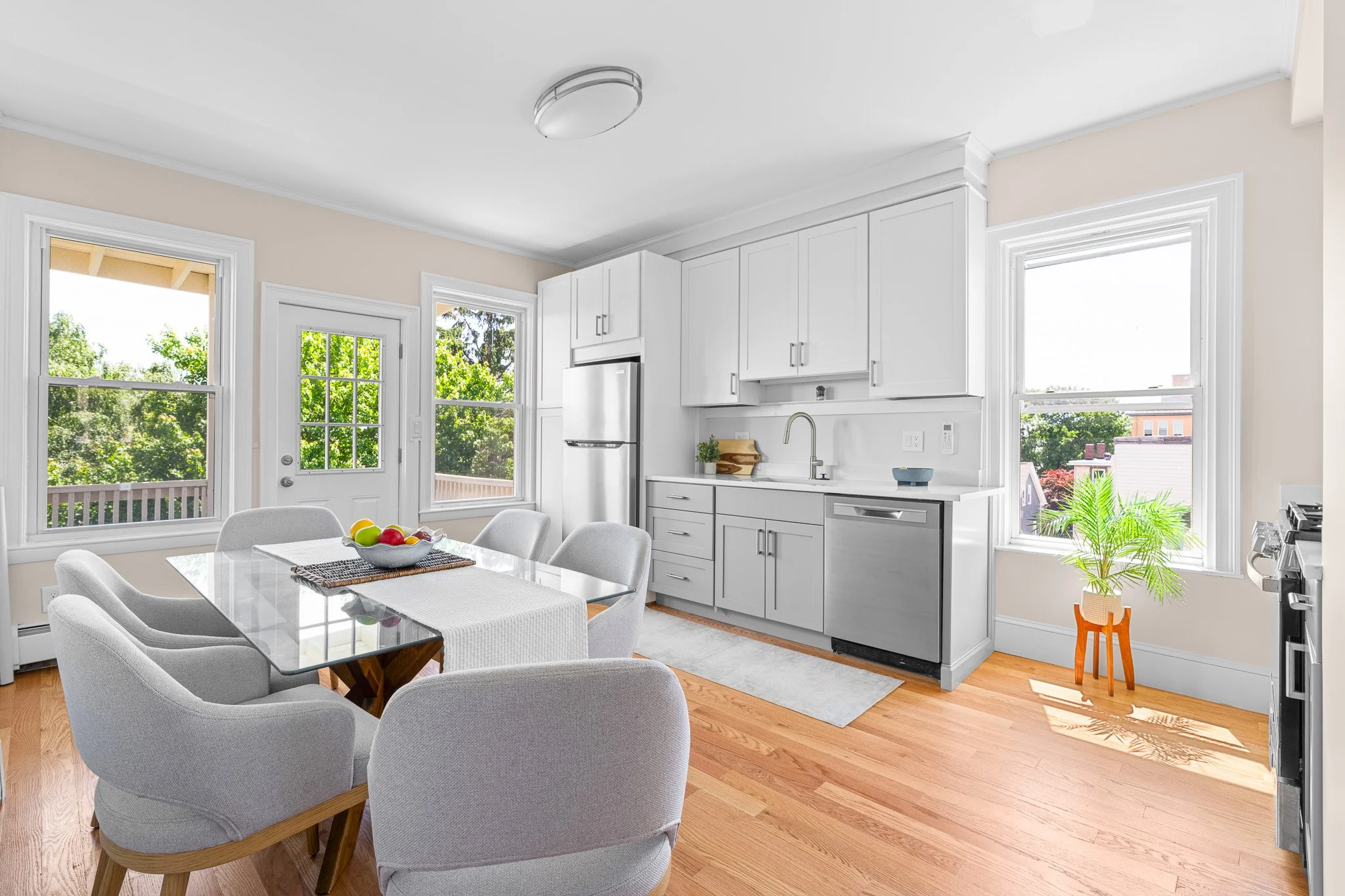 Bright kitchen and dining area with large windows, white cabinets, a dining table with chairs, a bowl of fruit, and a small plant by the window.