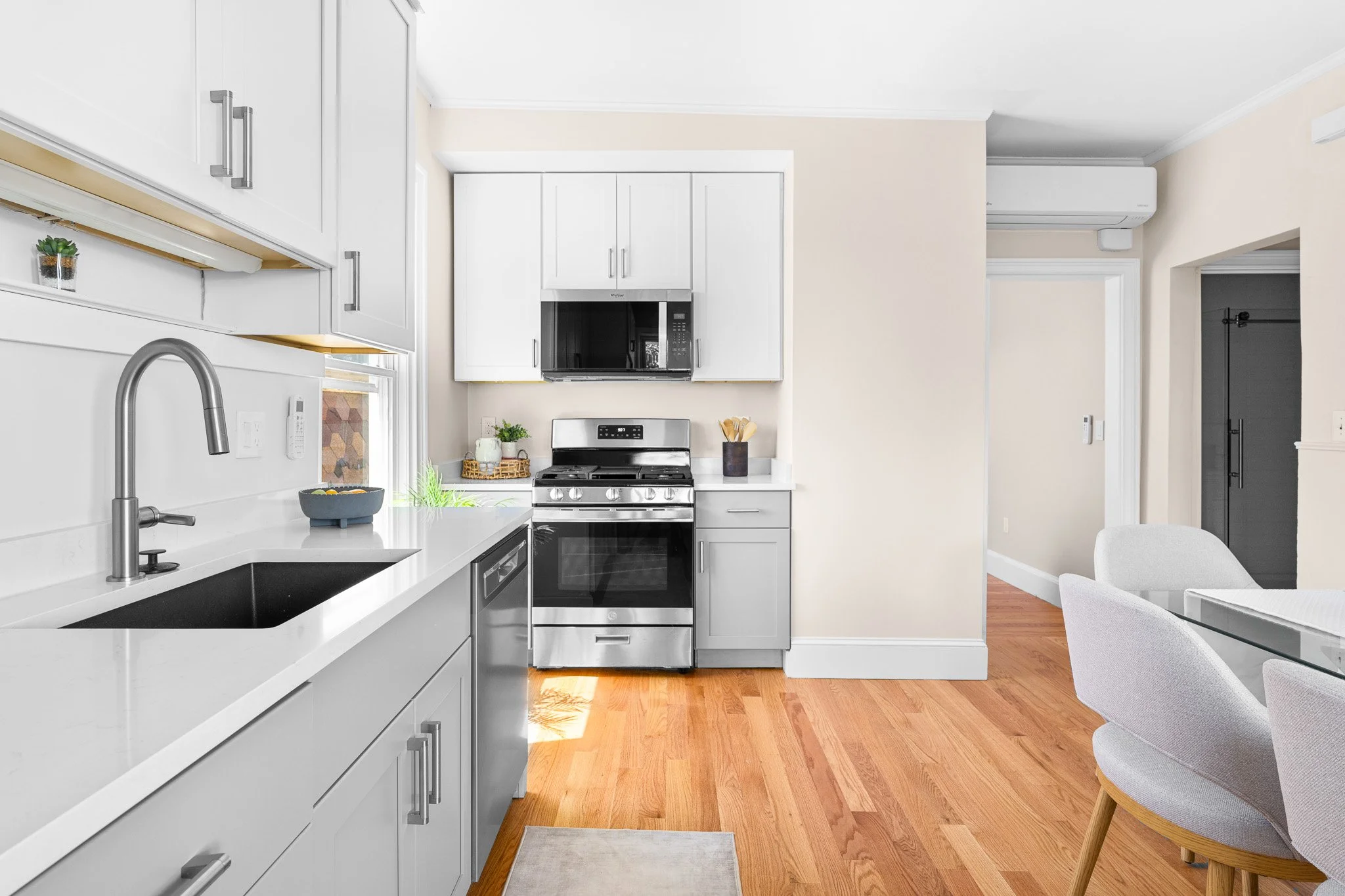 Modern kitchen with white cabinets, stainless steel appliances, a black sink, and a small dining area with a gray upholstered chair and a white table.