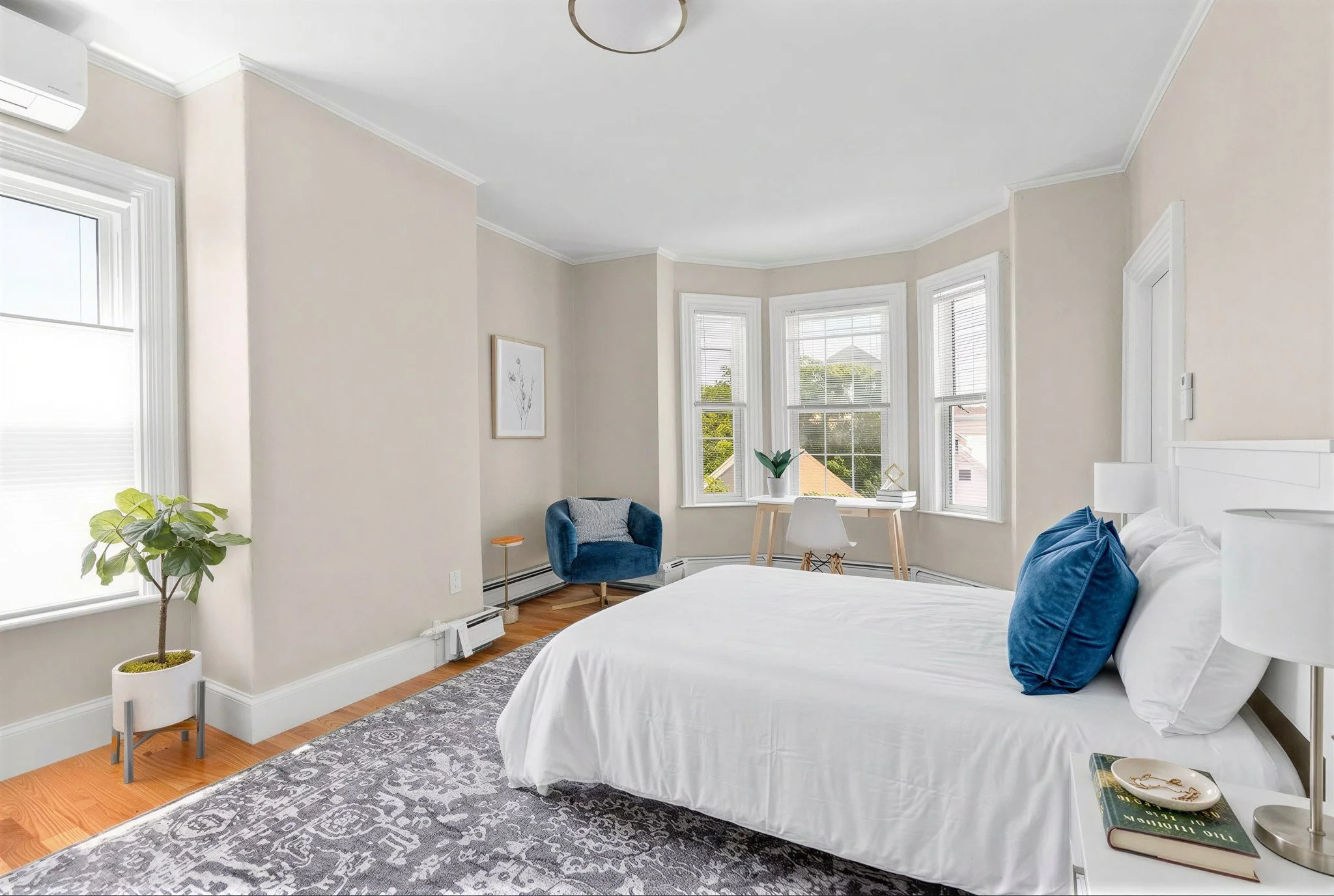 Bright bedroom with a white bed, blue pillows, and an adjacent nightstand holding books, a lamp, and a decorative dish. There is a large window with white blinds, a potted plant, a blue armchair, and a small side table near the window. The room has w