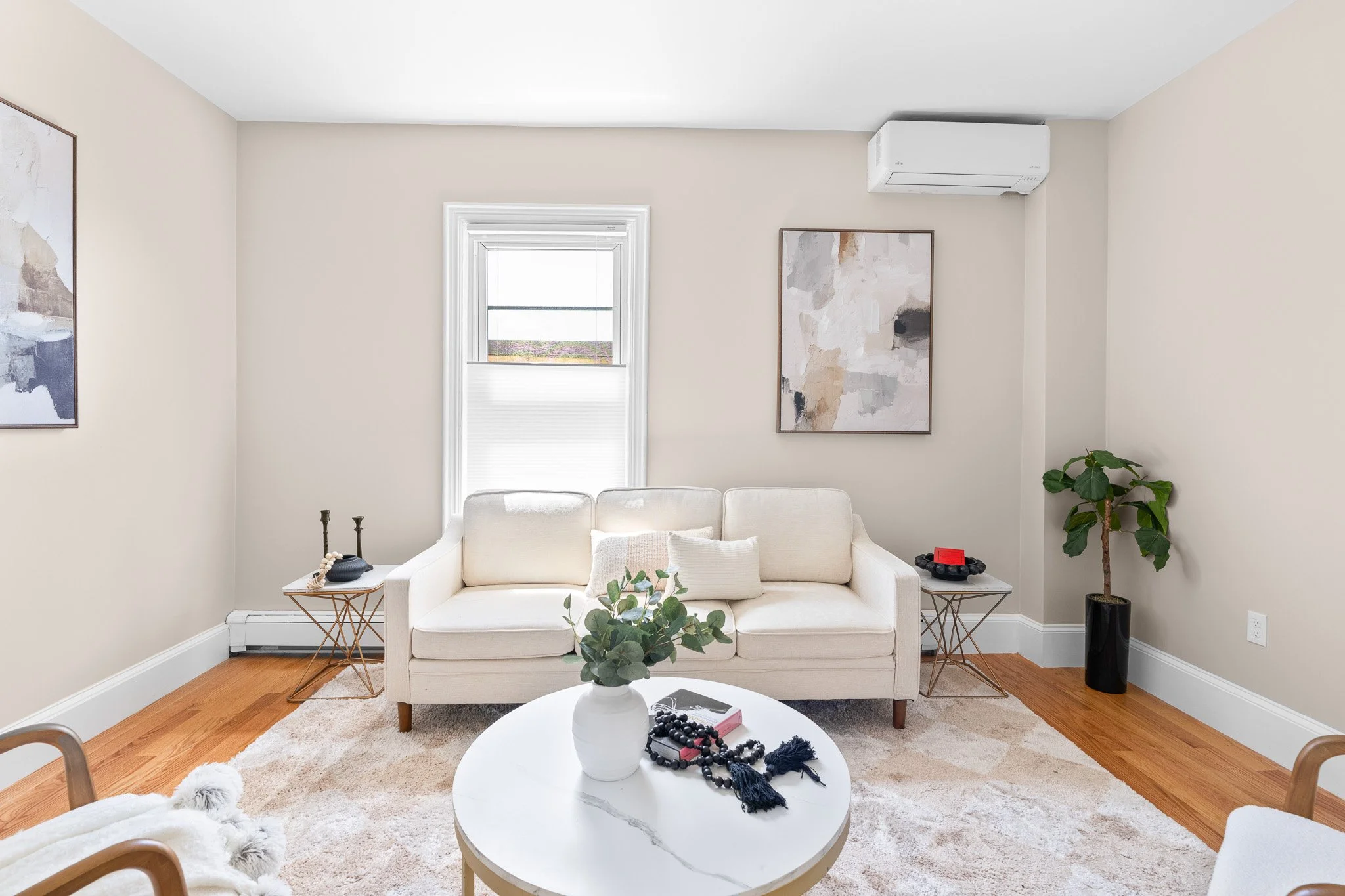 Living room with beige walls, white window, beige sofa with pillows, wood flooring, abstract artwork, coffee table with decorations, indoor plant, and air conditioner.